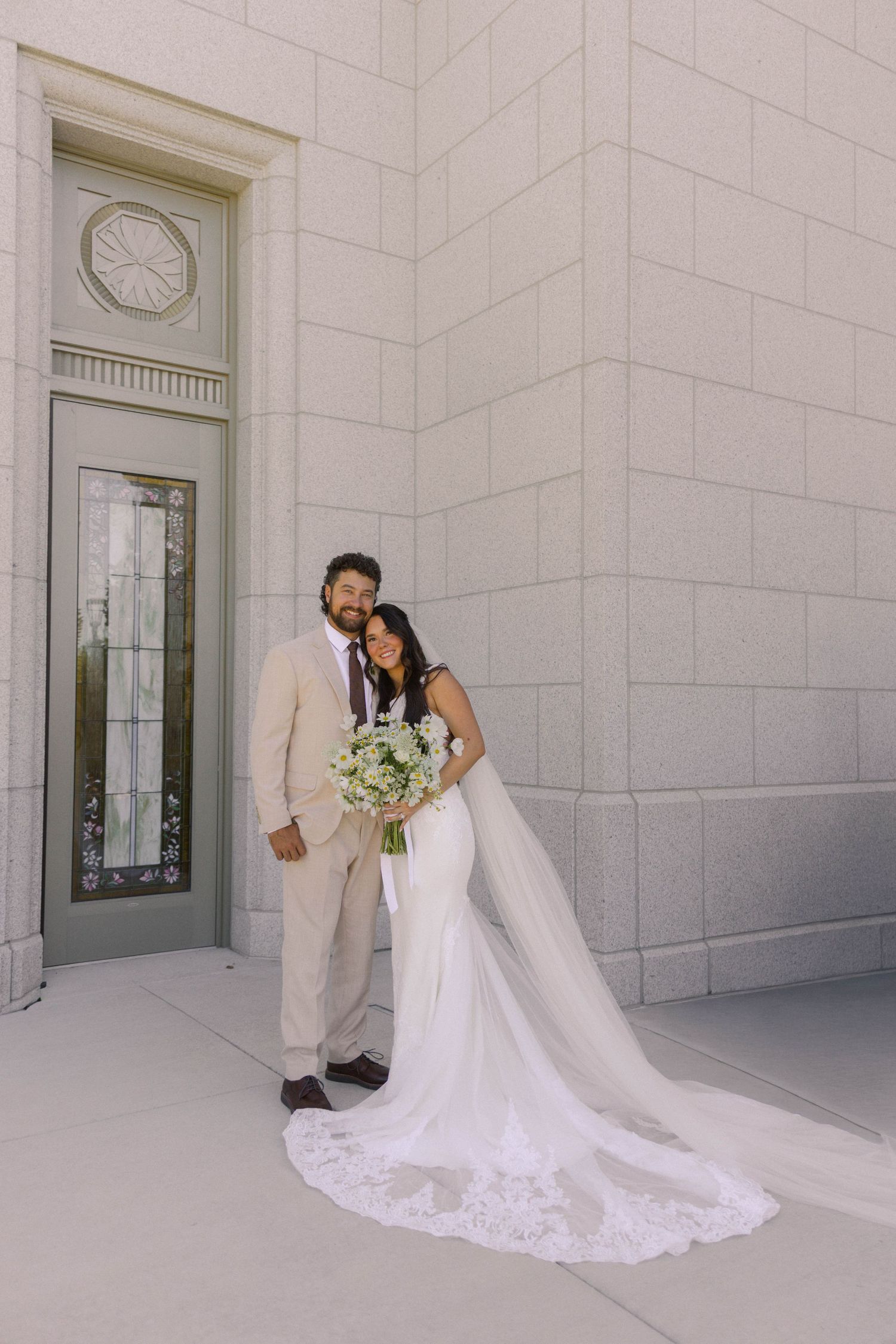 Bride in white lace gown with veil and groom in tan suit pose outside a stone building on their wedding day.