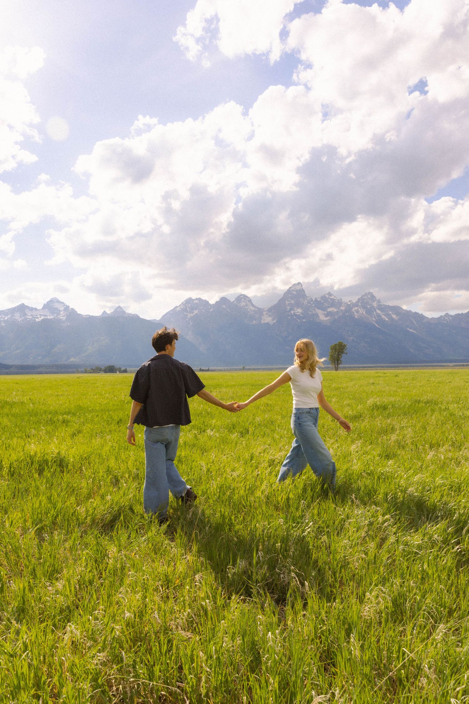 Couple holding hands walking through green meadow with Grand Teton mountains and dramatic clouds in background.