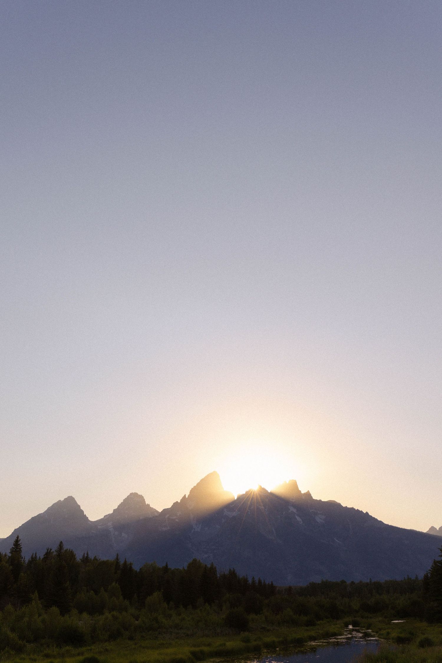 Sun setting behind jagged mountain peaks with evergreen forest and stream in Grand Teton National Park.
