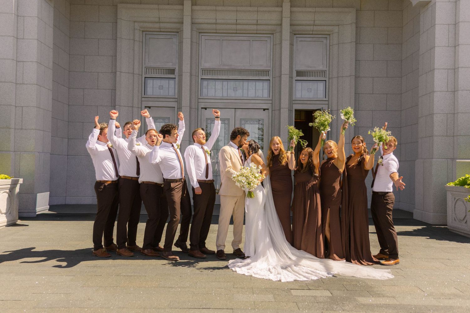 Wedding party celebrating outside a temple, bridesmaids in brown dresses and groomsmen in white shirts with suspenders cheering.