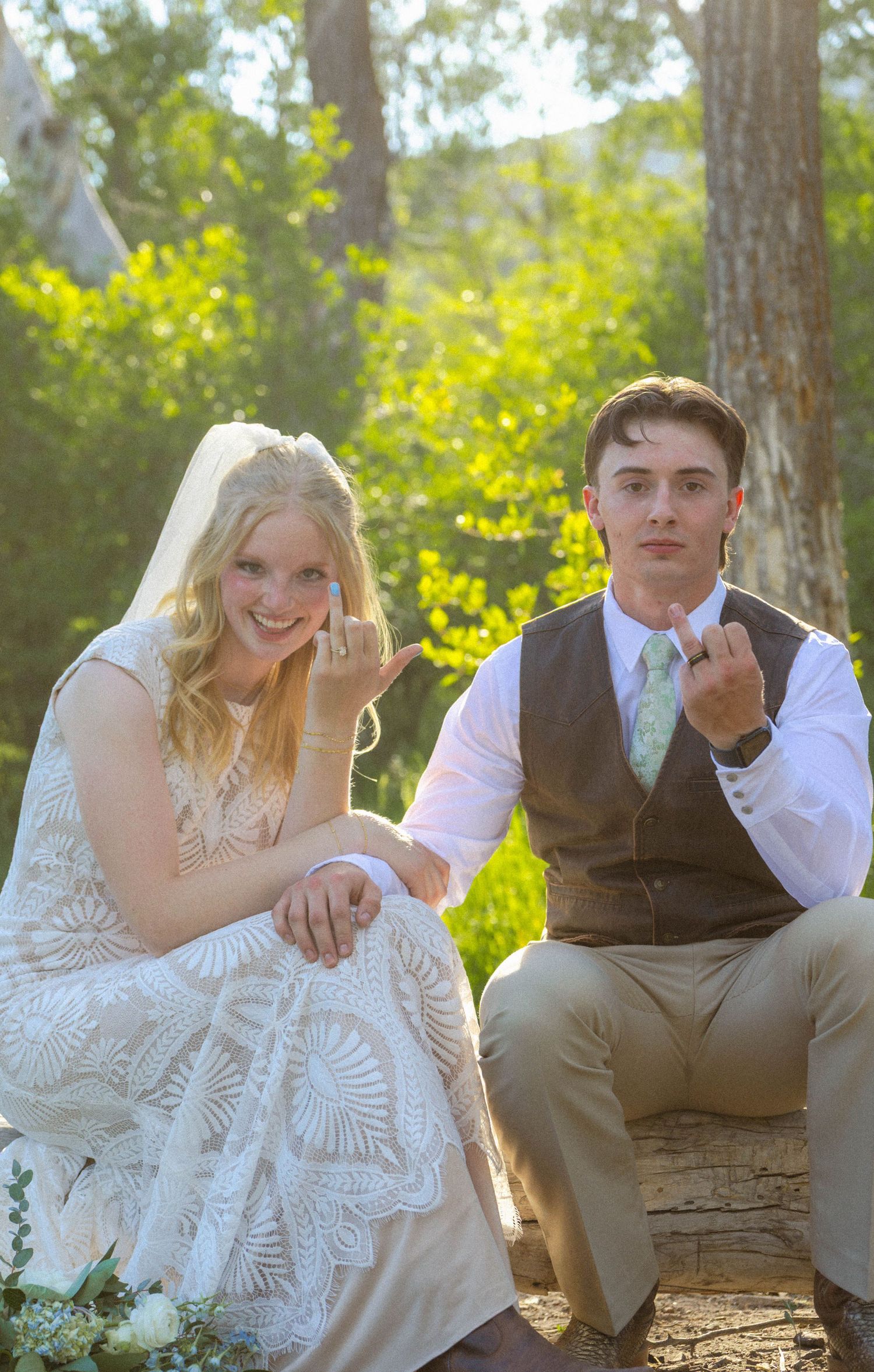 Bride and groom showing wedding rings outdoors, seated among green trees in wedding attire.