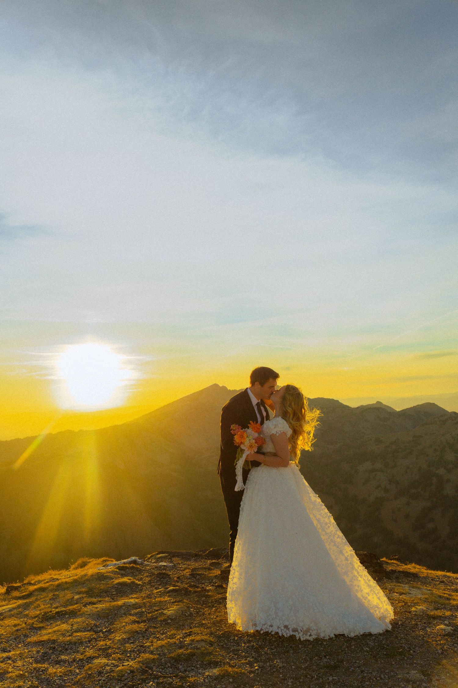 Bride and groom kissing at sunset on a mountain peak, with golden light and scenic hills in the background.