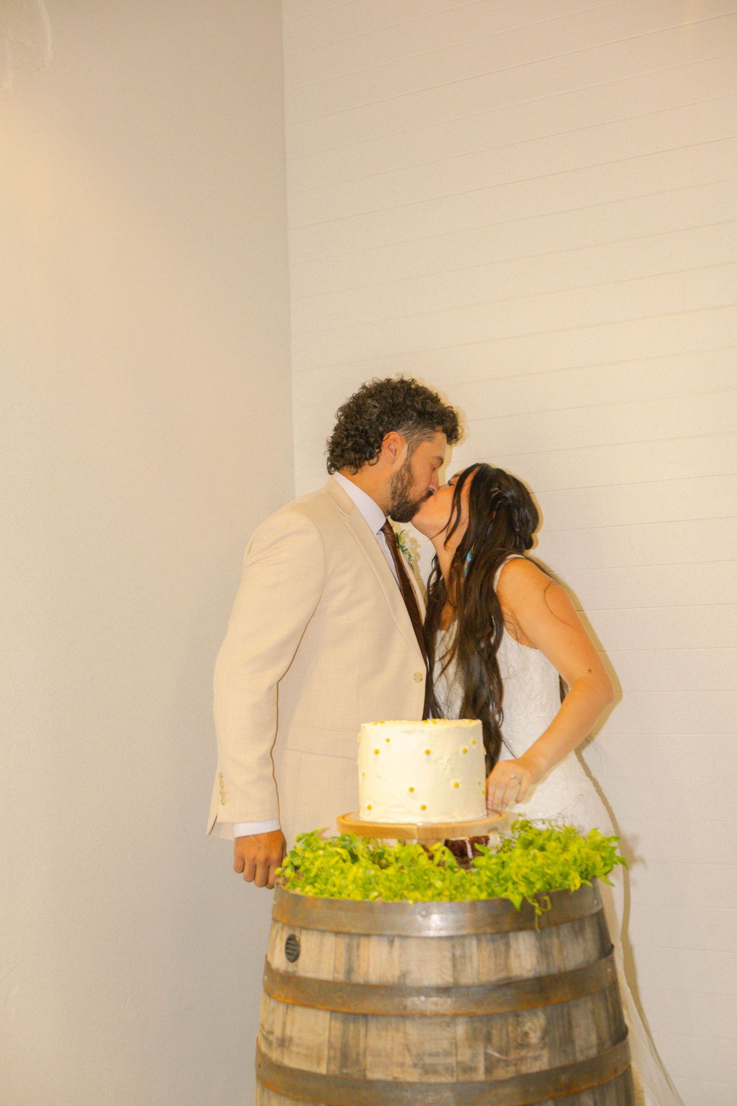 Bride and groom share a kiss while cutting their wedding cake displayed on a rustic wine barrel.