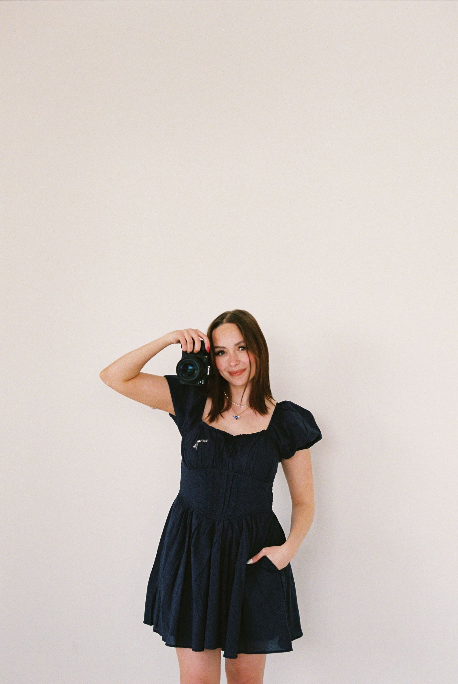 Young woman in black mini dress holding a camera, posing against a white wall in a stylish portrait photo.