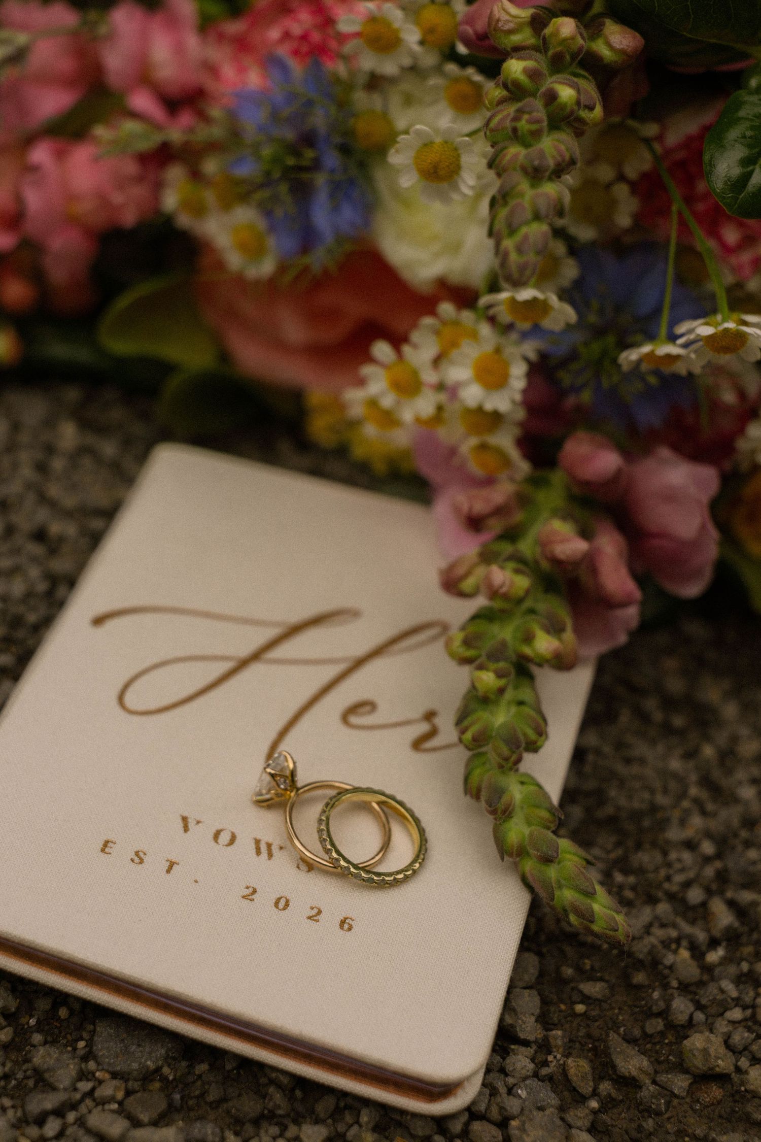 Gold wedding rings rest on a Her Vows Est. 2026 book beside a colorful floral bouquet with daisies and pink blooms.