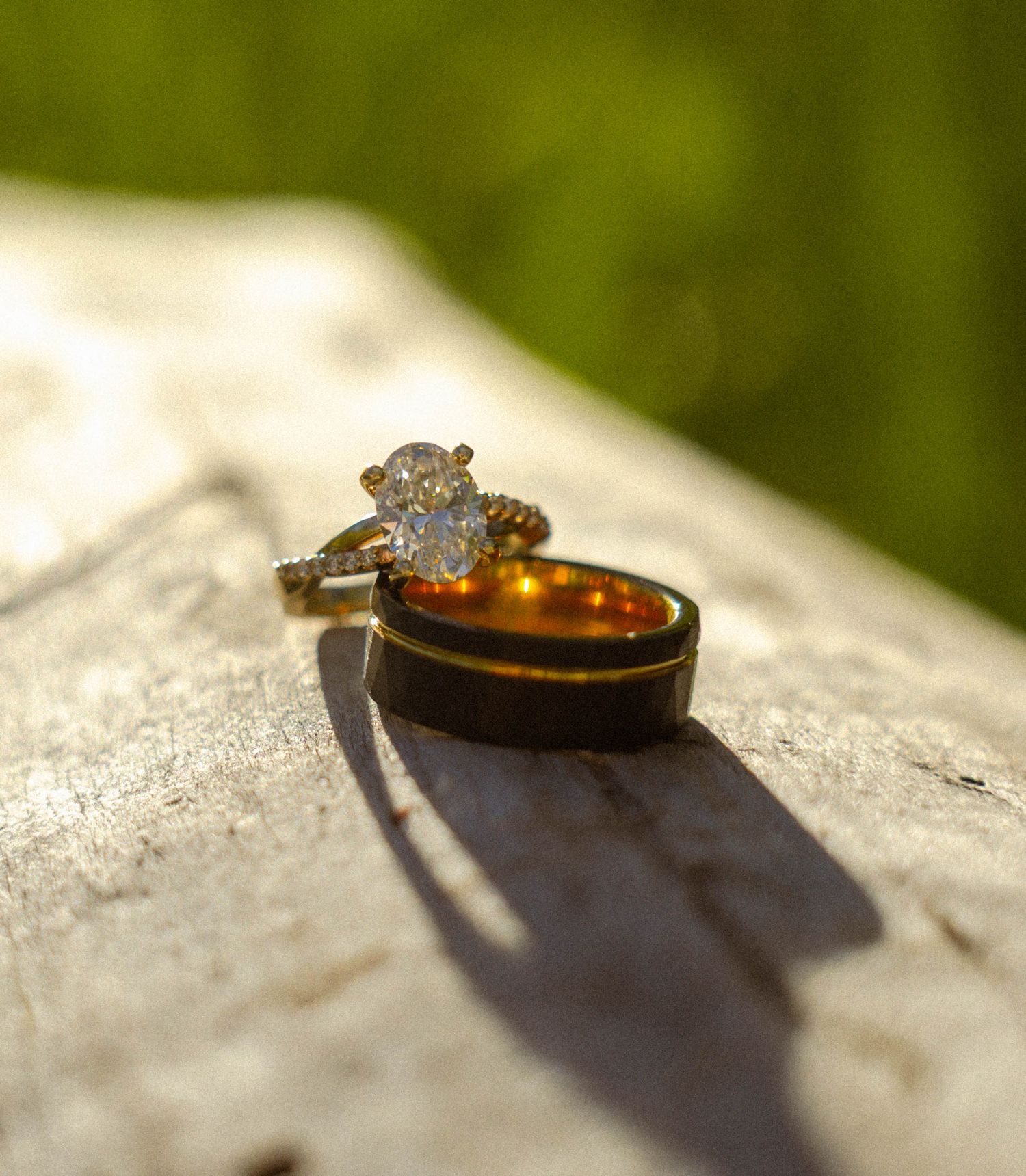 Gold diamond engagement ring and dark wood wedding band displayed on rustic wood surface with green bokeh background.