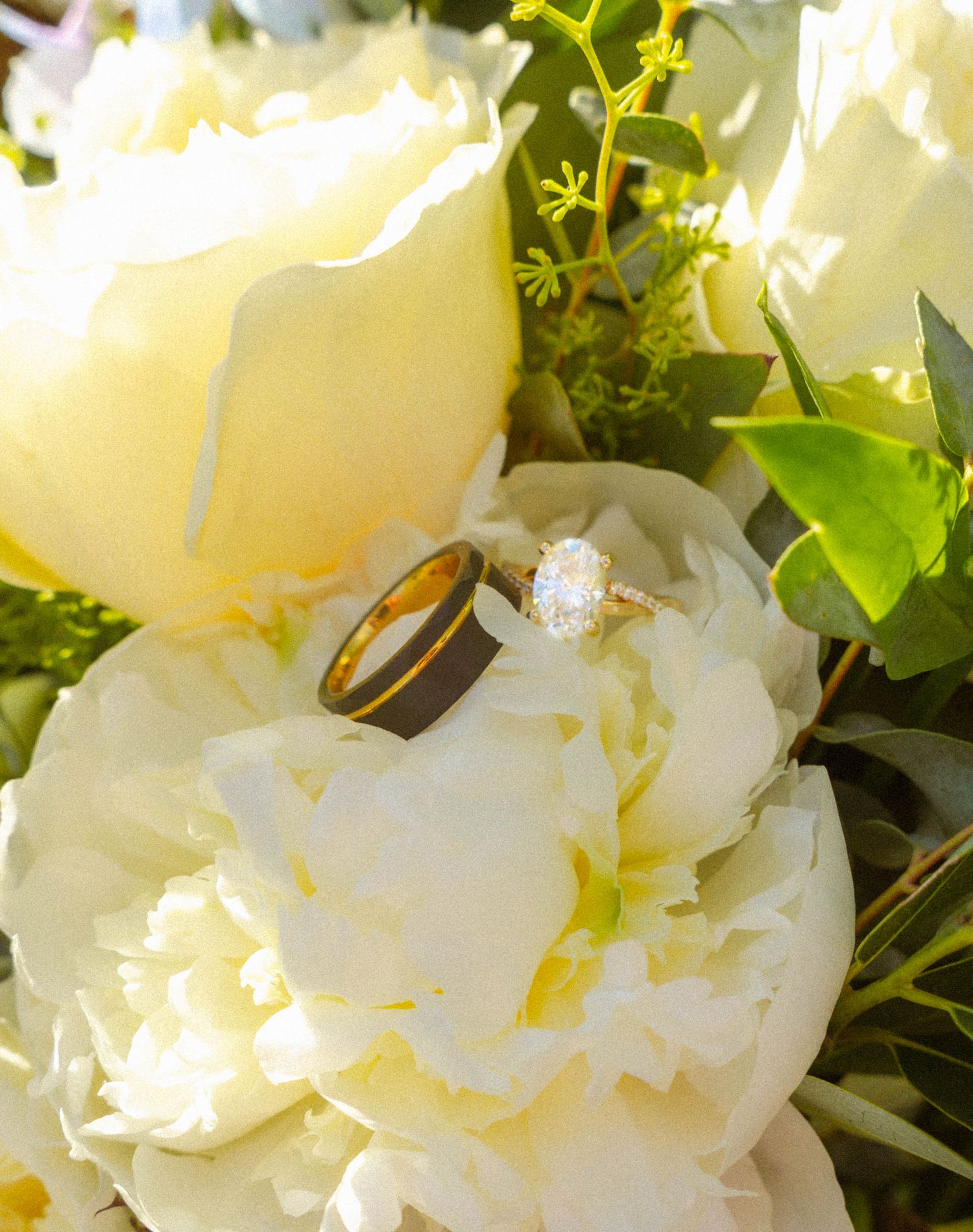 Gold engagement ring with diamond solitaire and wedding band resting on white peony flowers in a bridal bouquet.
