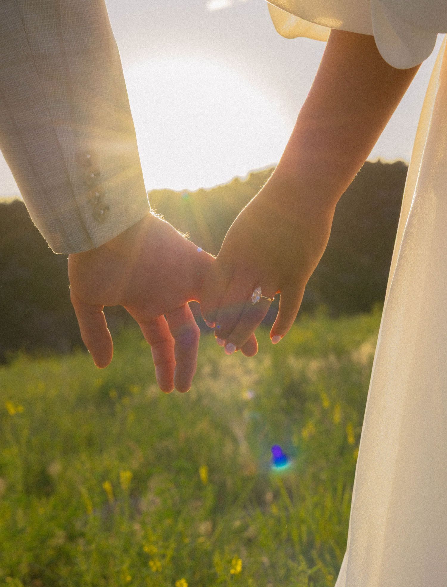 Couple holding hands in a sunlit field, wearing wedding attire with a diamond ring visible.