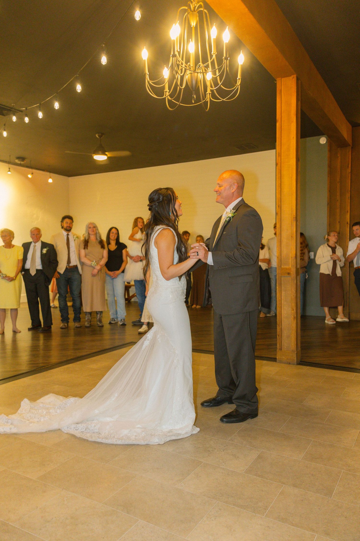 Bride and groom share their first dance in an elegant venue with chandelier lighting, guests watching in background.