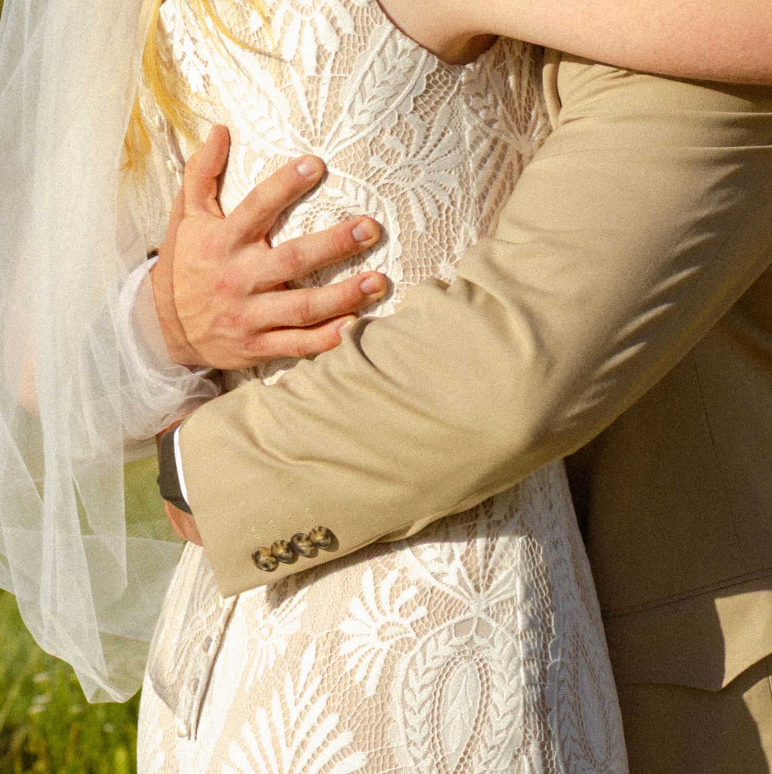 Bride and groom embracing, showcasing lace wedding dress and tan suit with wedding rings visible.
