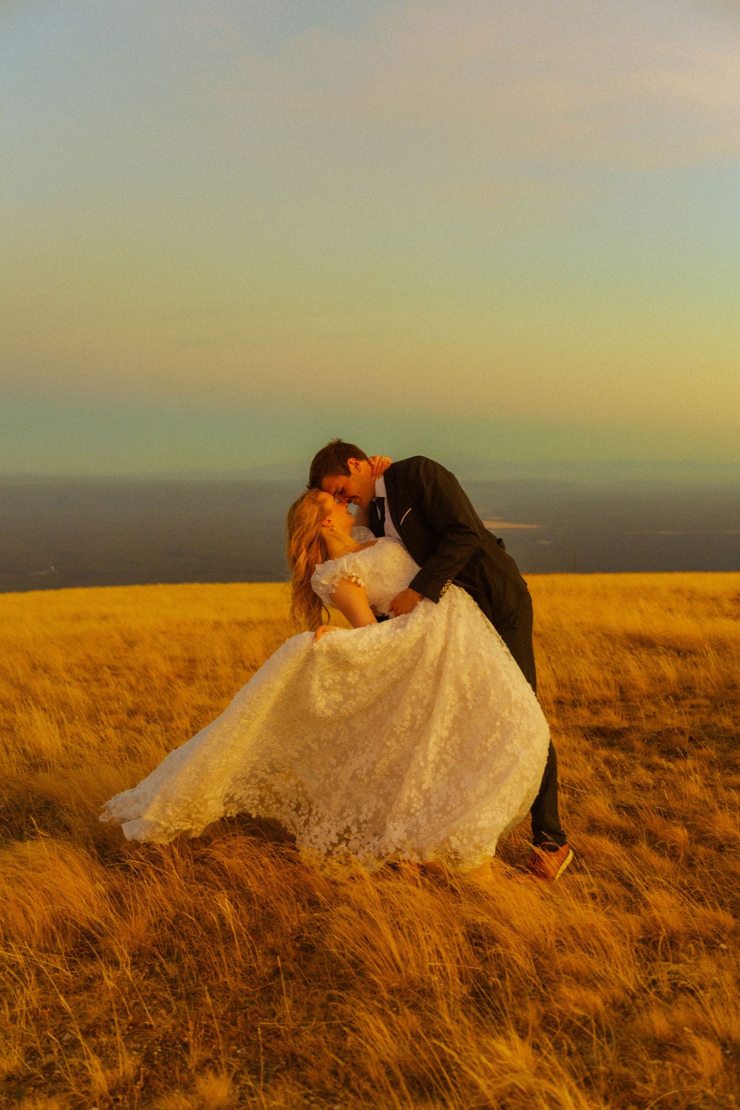 Bride and groom sharing a romantic kiss in a golden field at sunset, with a sweeping landscape in the background.