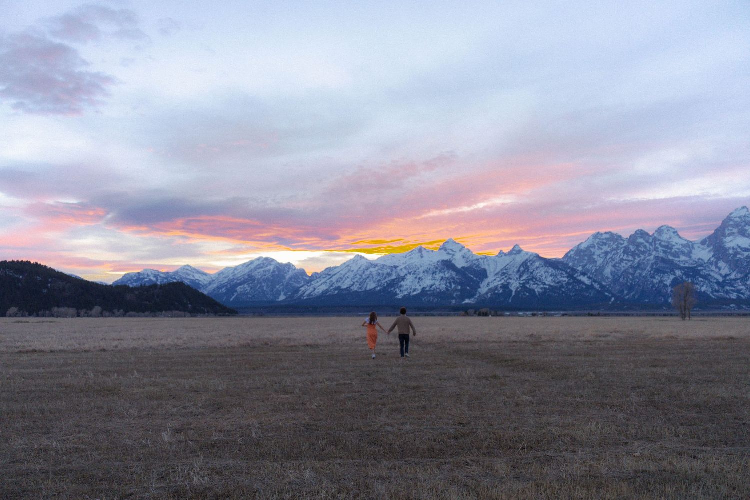 Couple walking hand-in-hand across open field toward colorful sunset and snow-covered Grand Teton mountains.