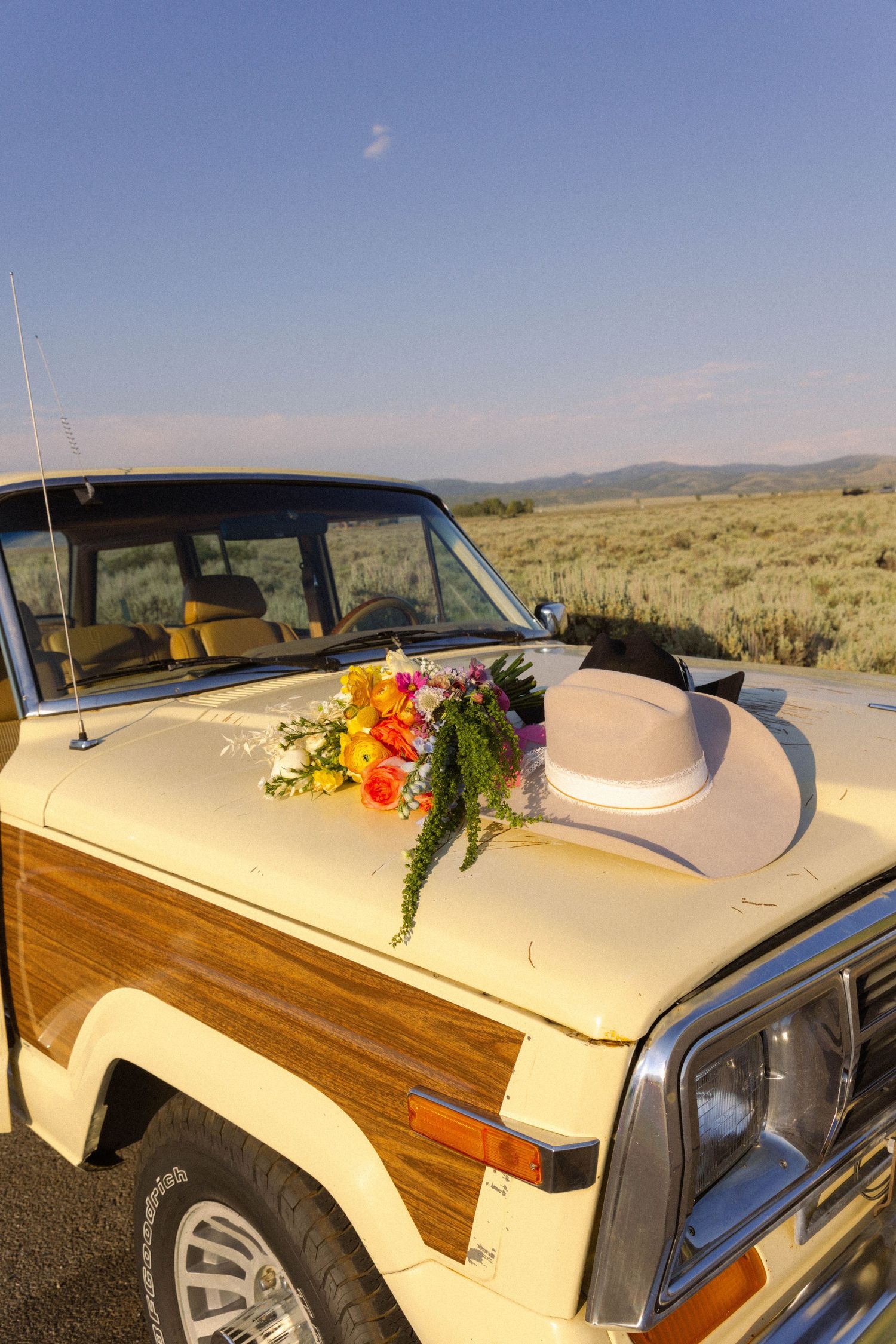 Vintage Jeep Wagoneer with wood paneling, topped with colorful flowers and white hat, parked in open sagebrush landscape.