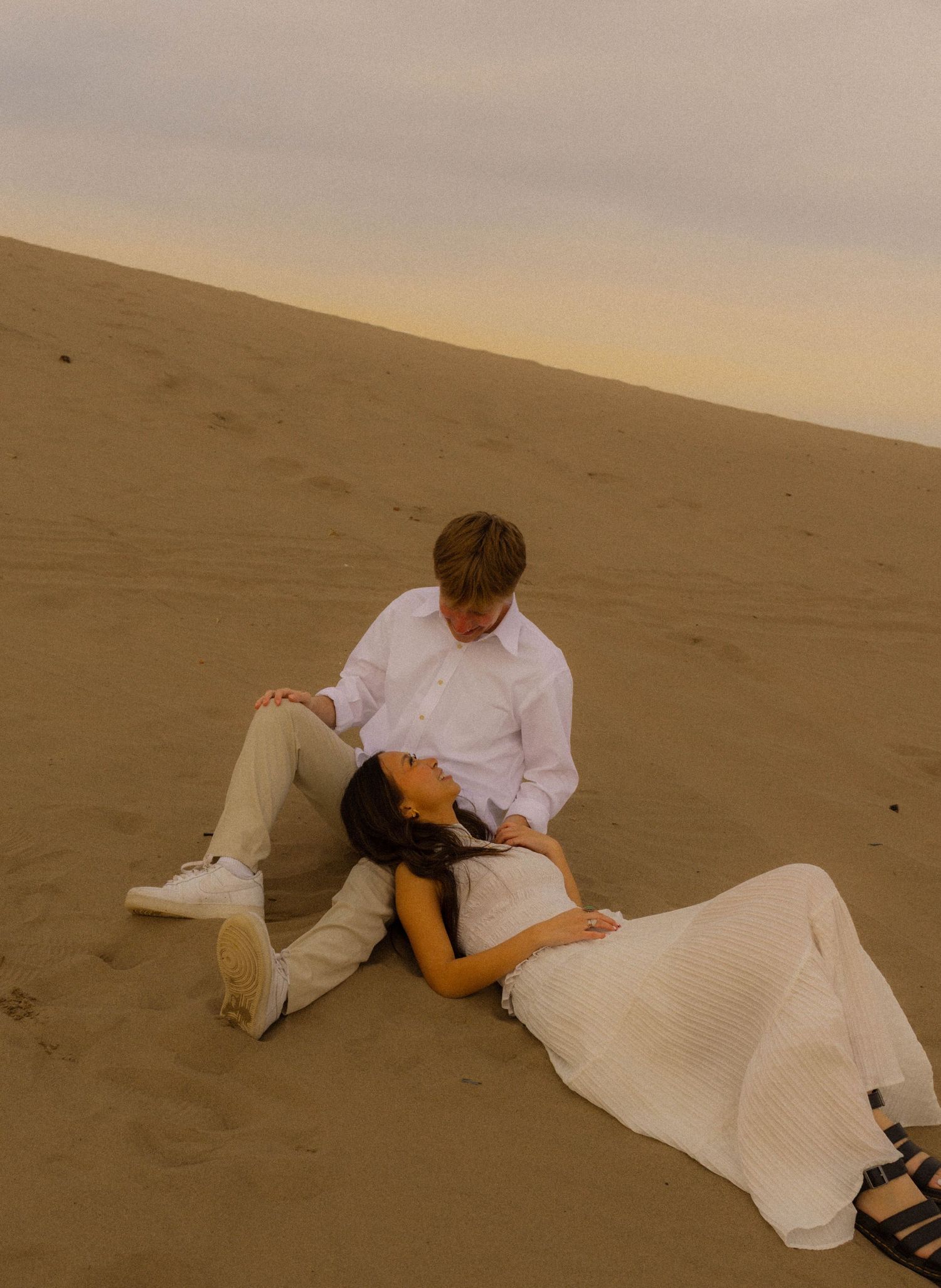 Couple in white outfits relaxing on sandy dunes at sunset, woman lying across man's lap romantically.