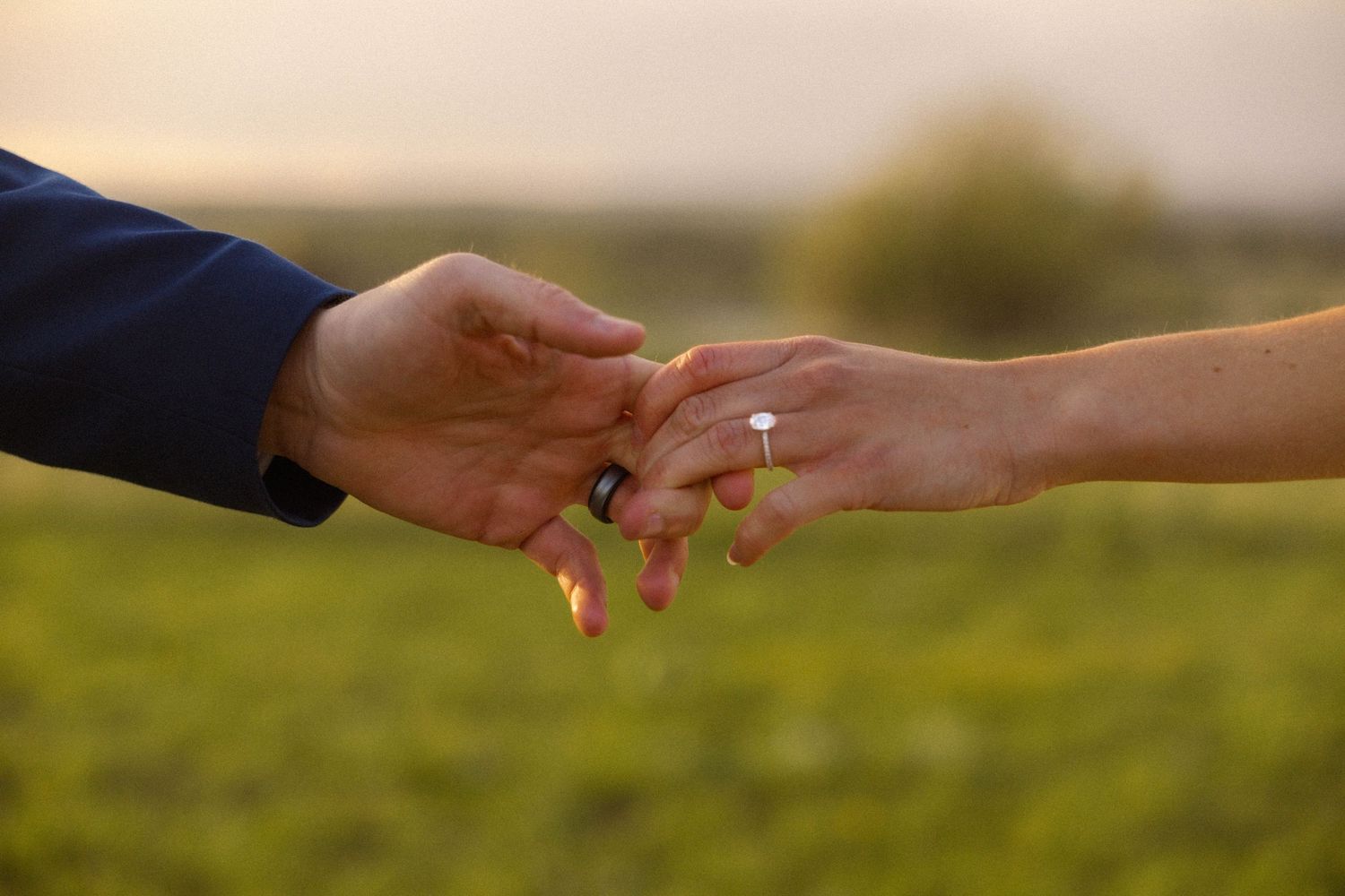 Couple holding hands outdoors at sunset, woman wearing a diamond engagement ring, green field in background.