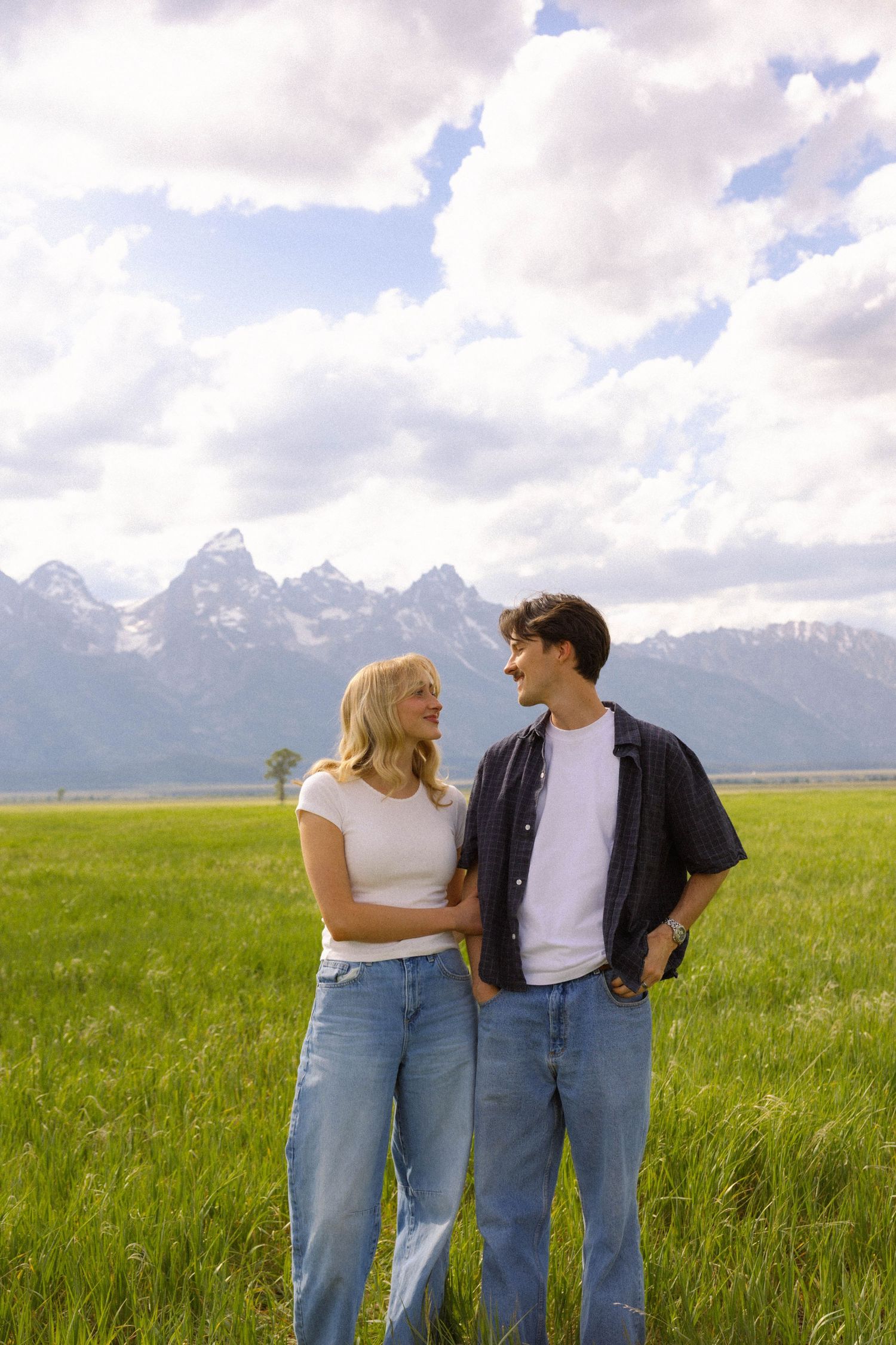 A couple stands in a green meadow with the Grand Teton mountains and cloudy sky behind them.
