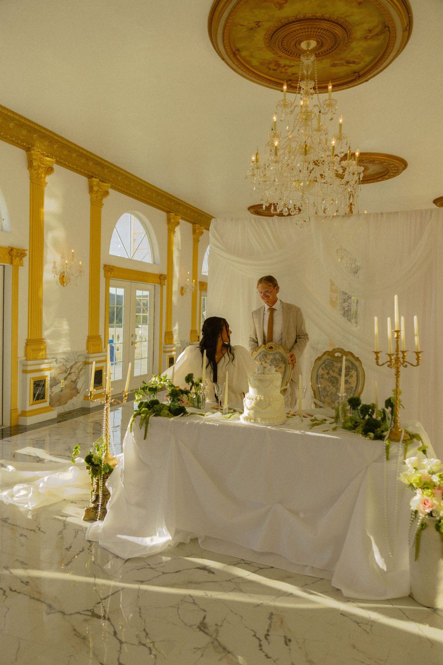 Elegant wedding reception in a grand ballroom with gold accents, chandelier, marble floors, and couple at a floral sweetheart table.