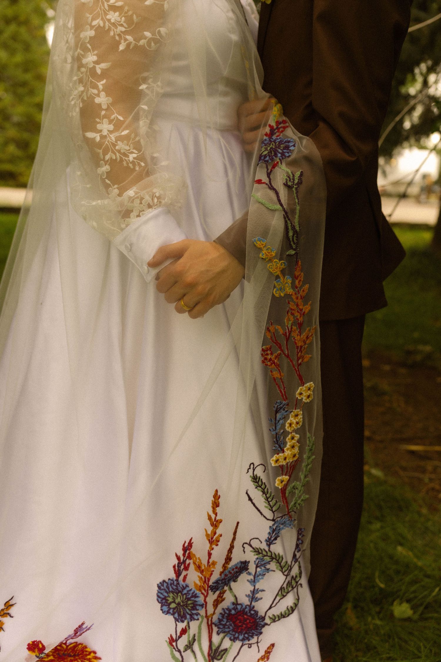 Bride in white gown with colorful floral embroidered veil stands with groom in brown suit outdoors.