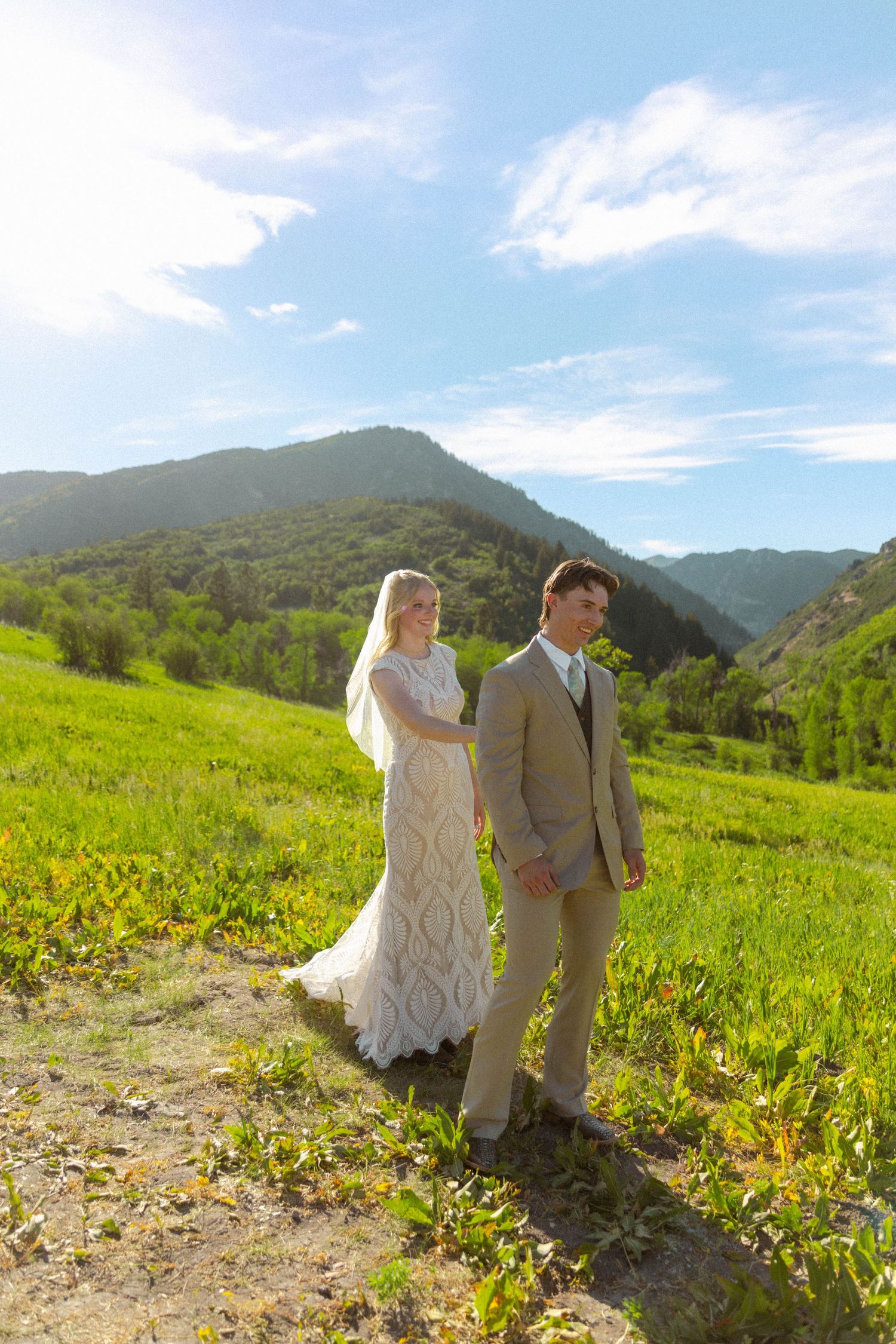 Bride and groom holding hands in a lush green mountain meadow under a bright blue sky on their wedding day.