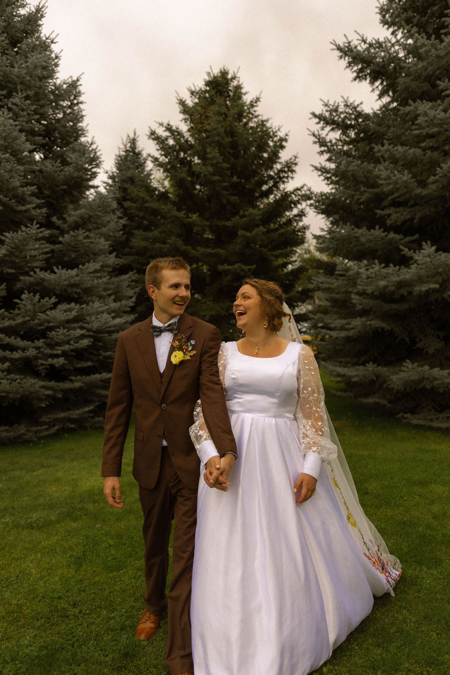 Happy bride and groom walking hand in hand on green grass with tall evergreen trees in background.