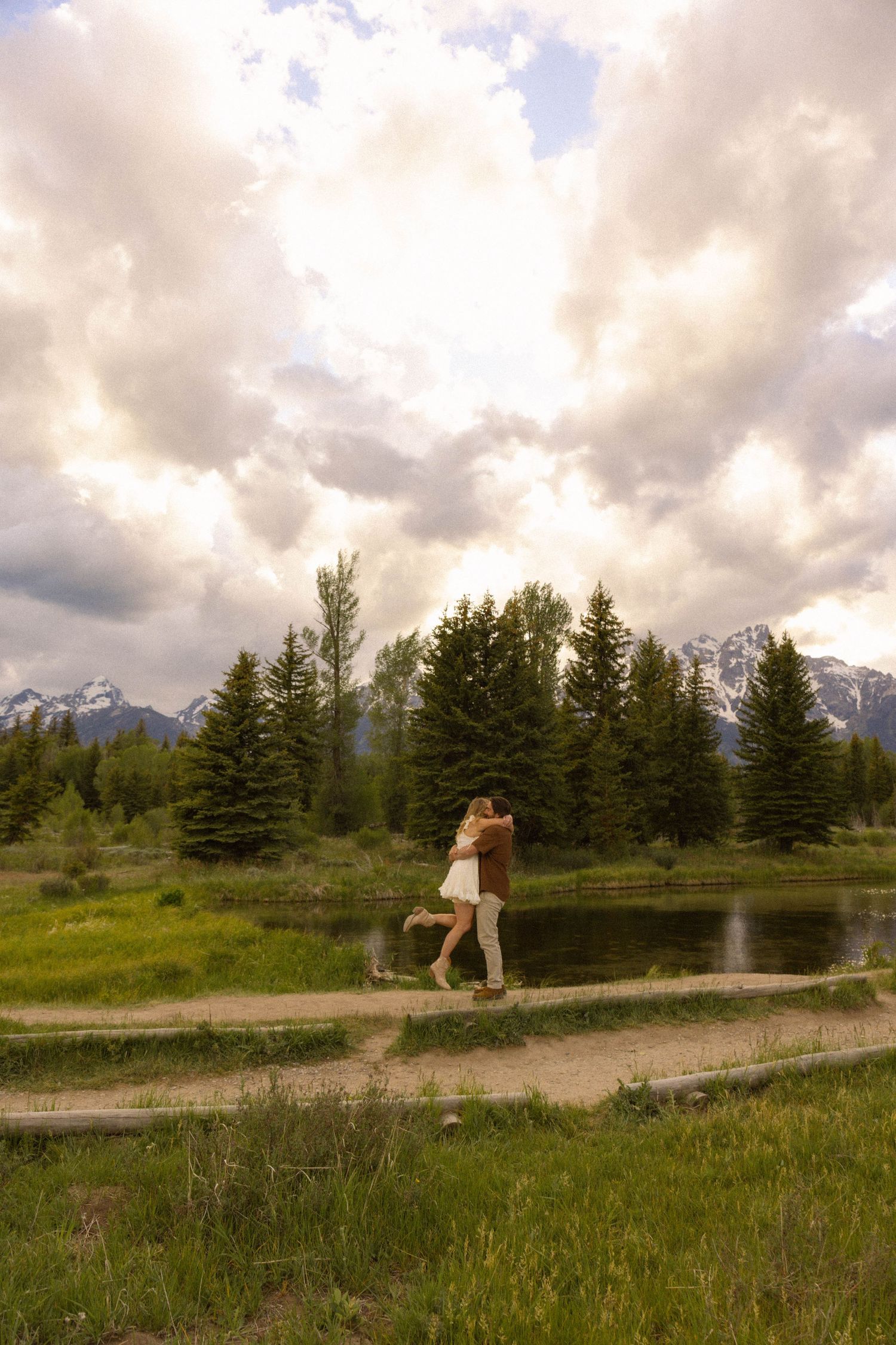 Couple embracing on a path near a pond with evergreen trees and snow-capped mountains under dramatic cloudy skies.