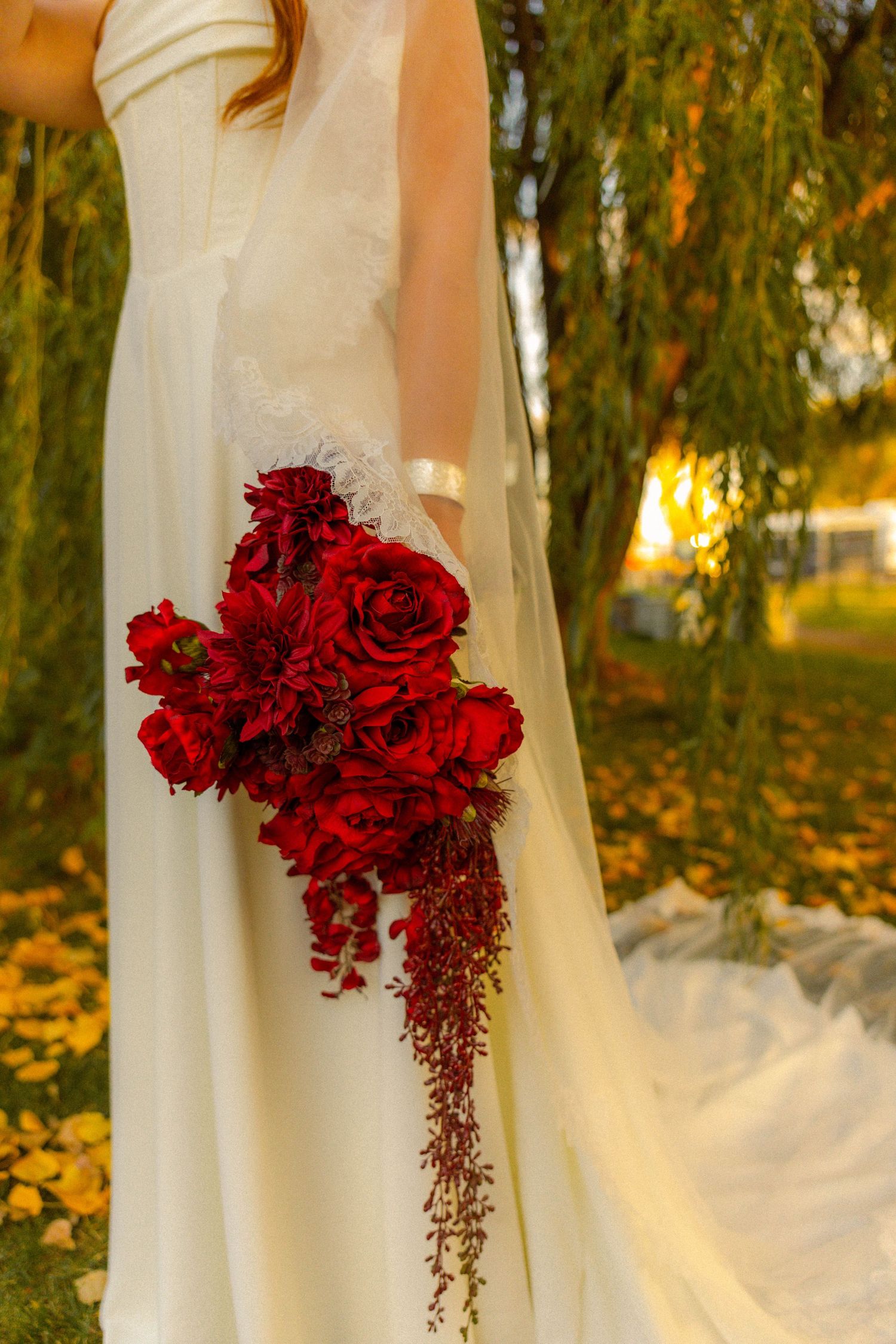 Bride in white gown holding cascading red rose and dahlia bouquet outdoors among fall foliage.