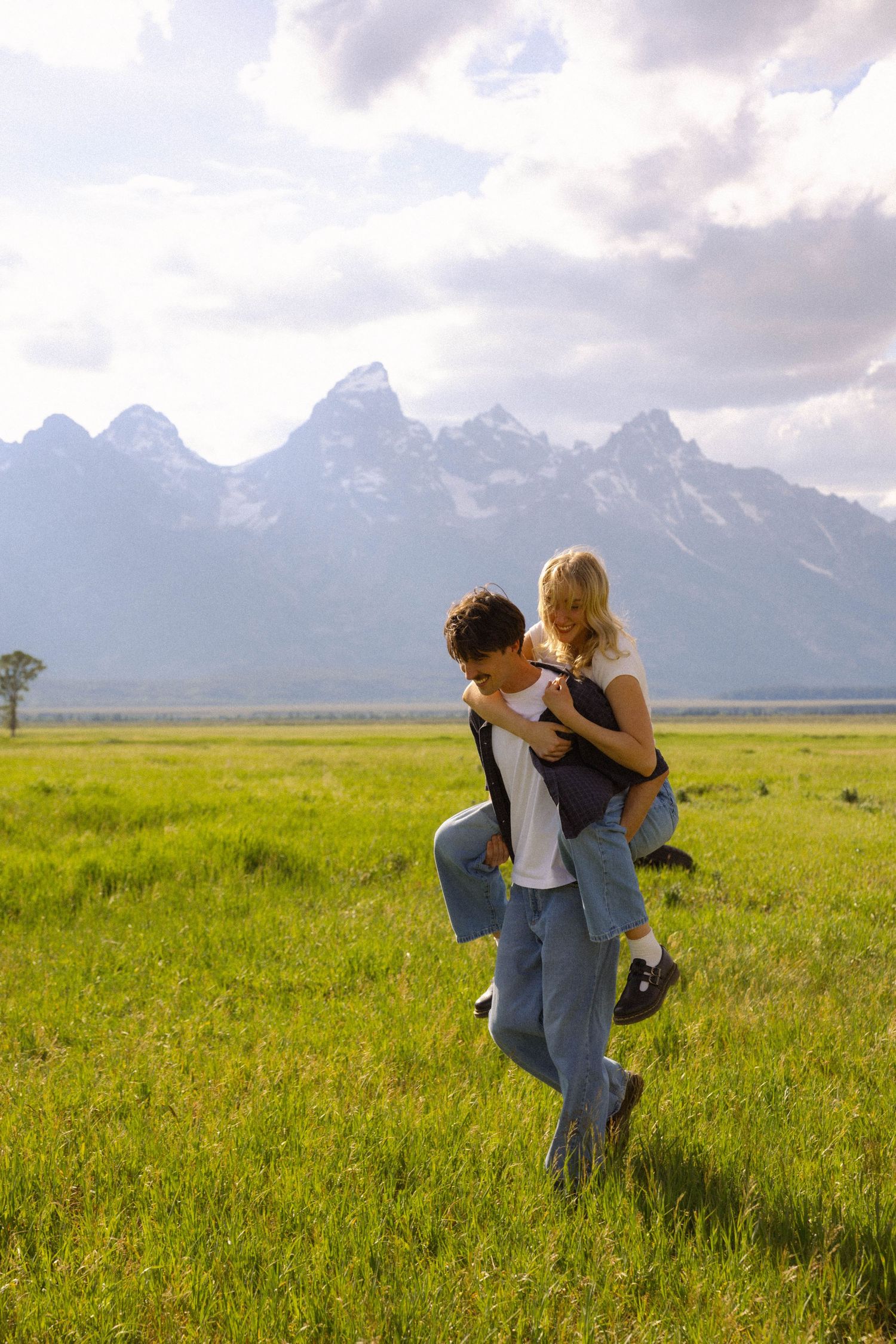 Couple sharing a piggyback ride in a green meadow with Grand Teton mountain range in the background.