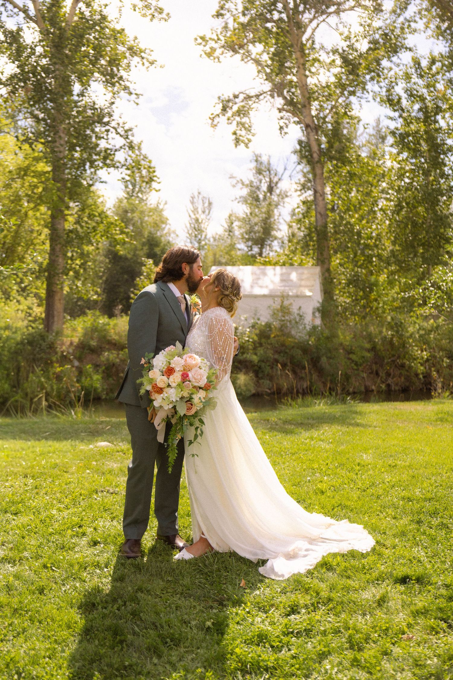 Bride and groom sharing a kiss outdoors on green grass, surrounded by tall trees, bride holding a pink and white bouquet.