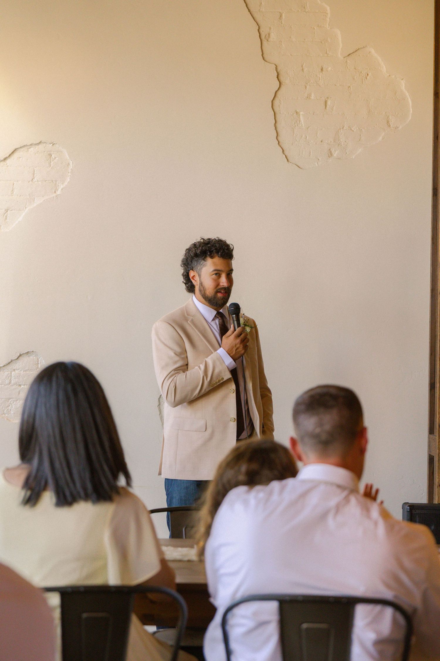Man in beige blazer speaking into microphone before seated audience in bright modern venue.