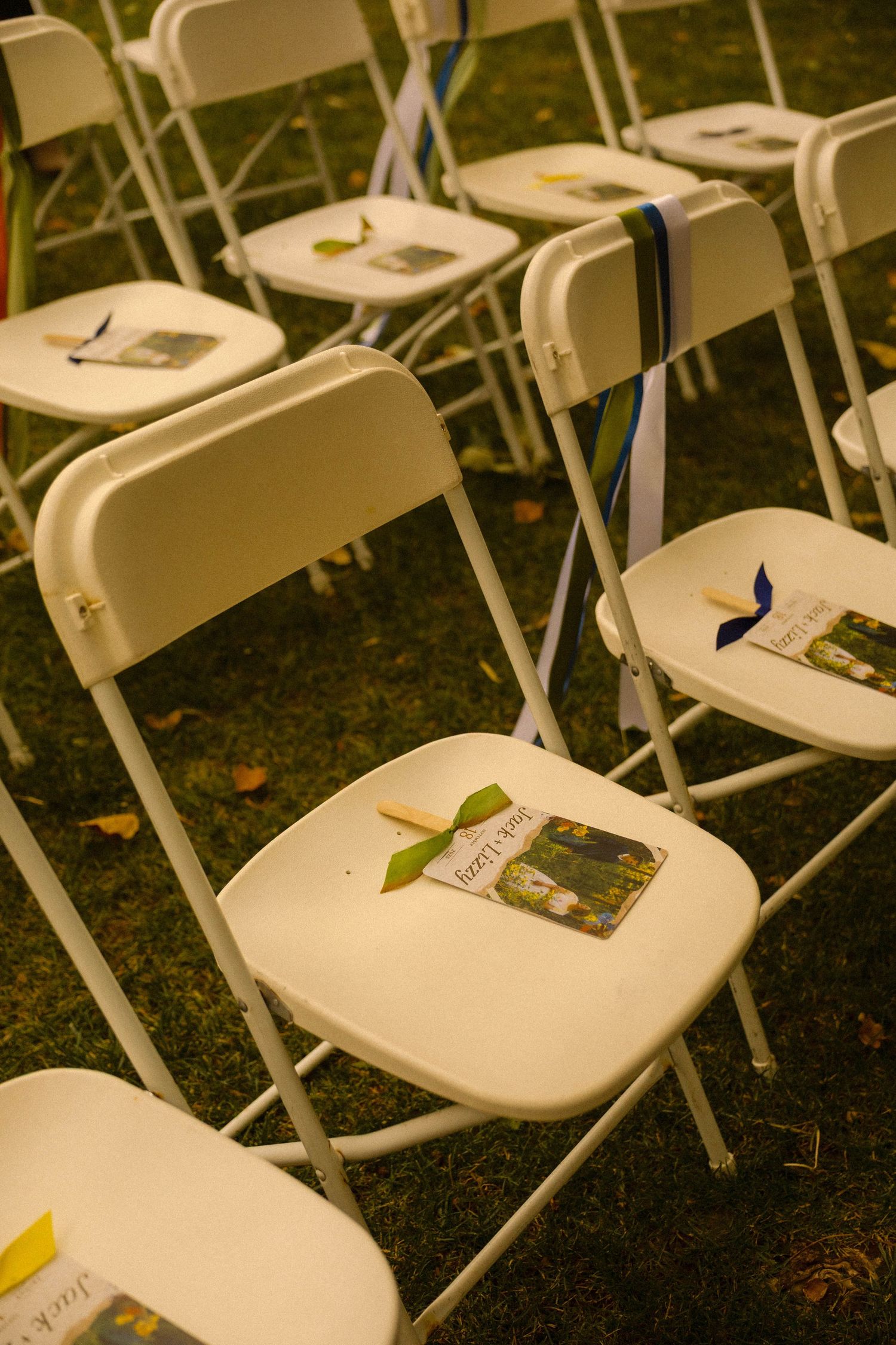 White folding chairs on grass with programs and ribbons for an outdoor wedding ceremony.