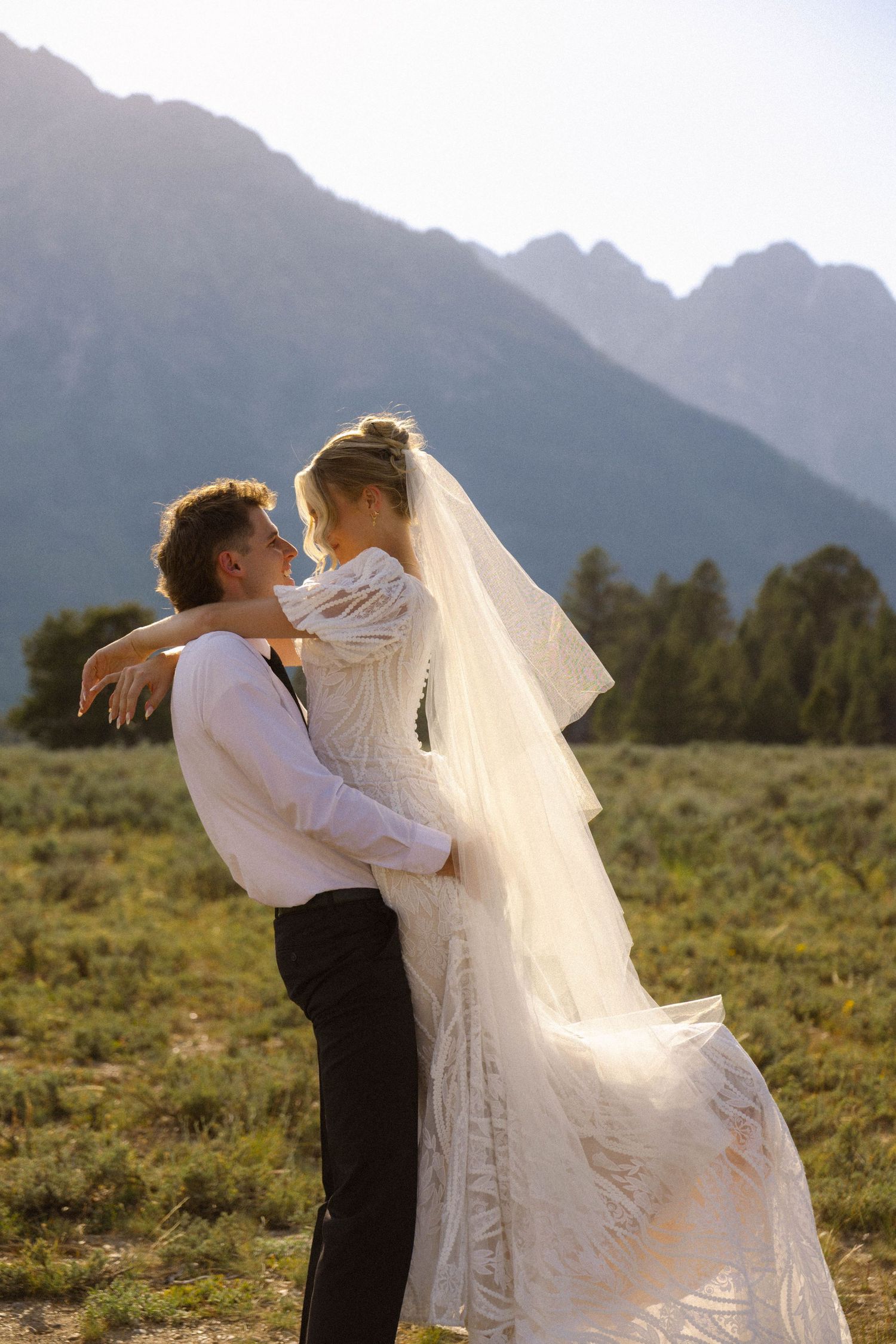 Bride and groom embrace in a sunlit mountain meadow, she wears a lace gown and veil.