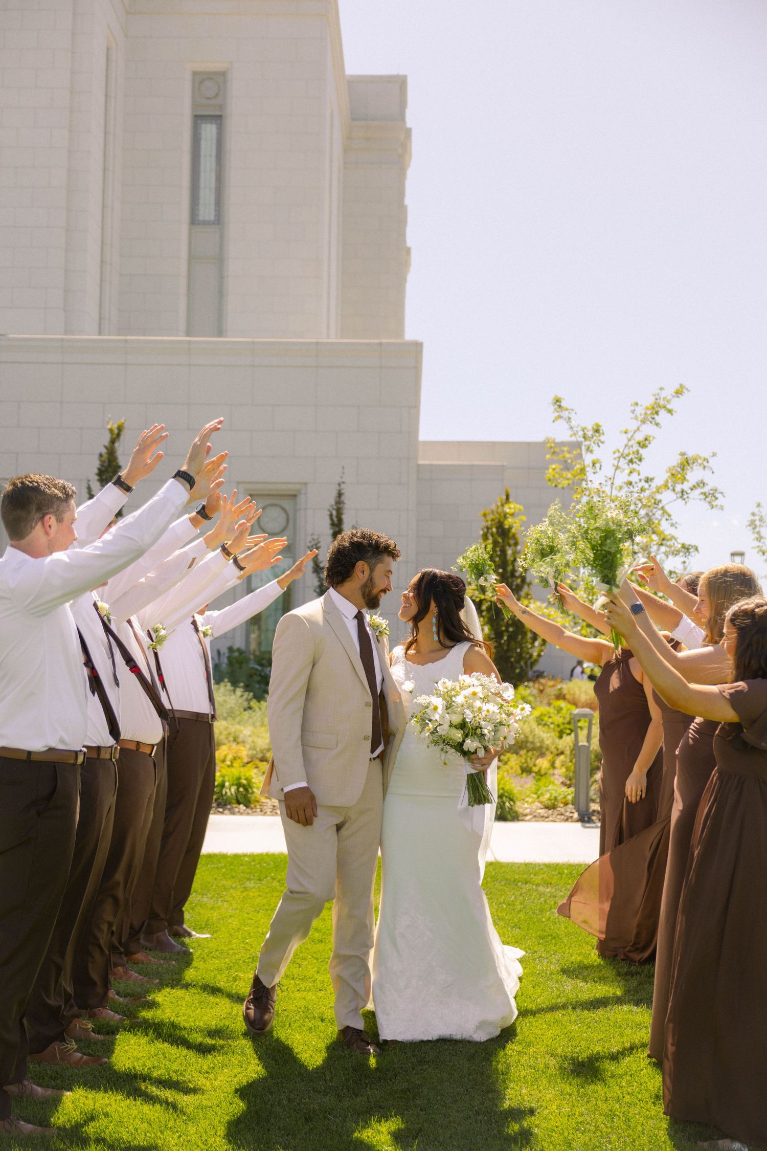 Bride and groom walk through arch of raised hands formed by bridesmaids in brown dresses and groomsmen outside white temple.