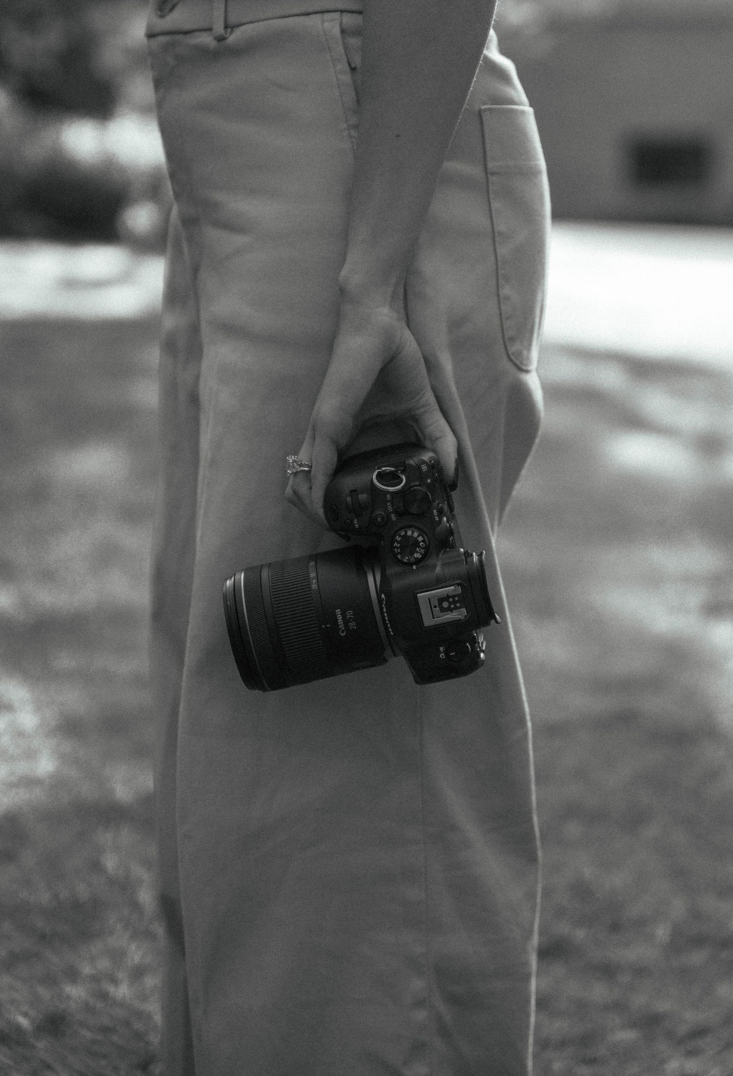 Black and white photo of a person holding a DSLR camera at their side while standing outdoors.