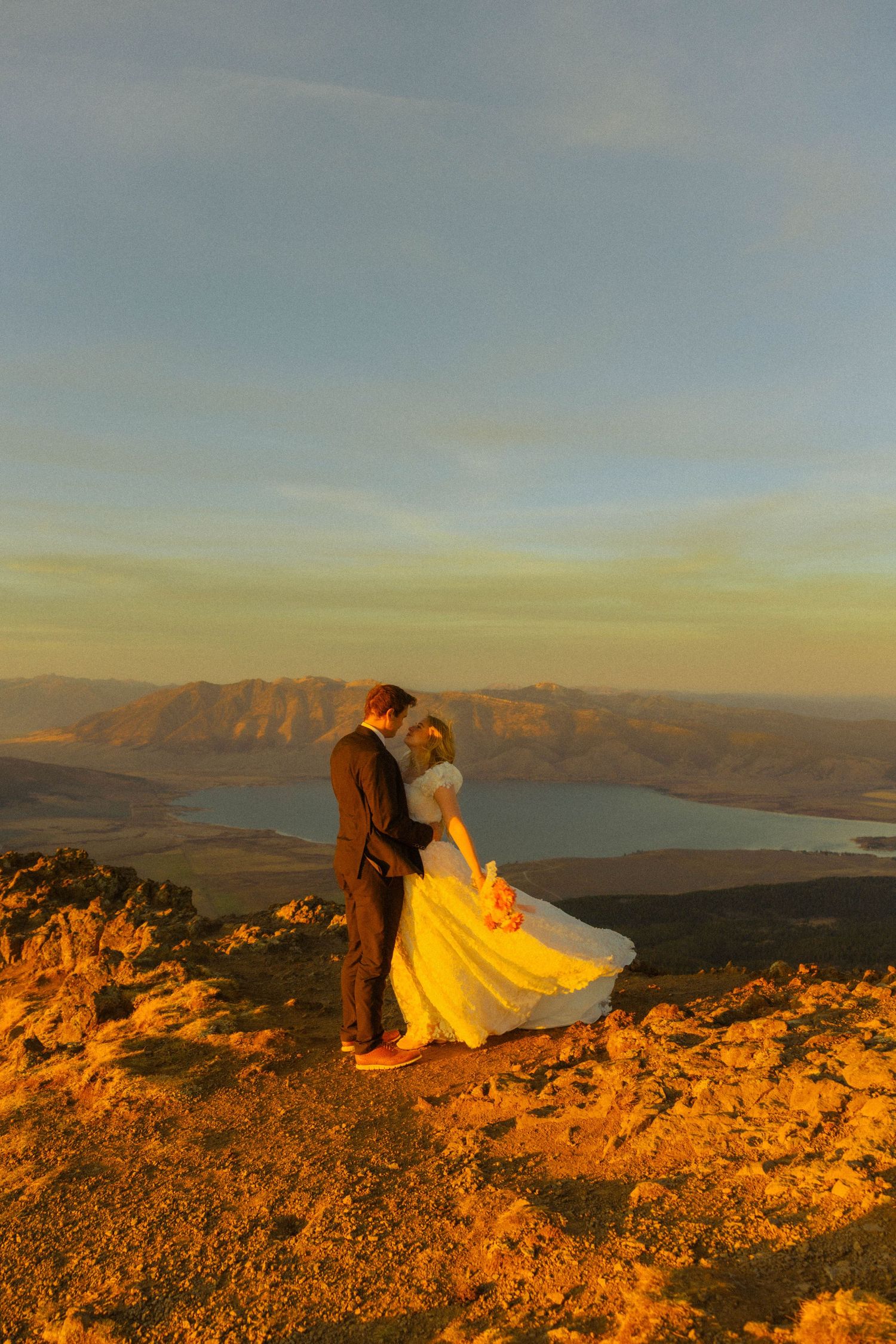 Couple embracing in wedding attire on mountain summit at golden sunset with lake and mountain range backdrop.