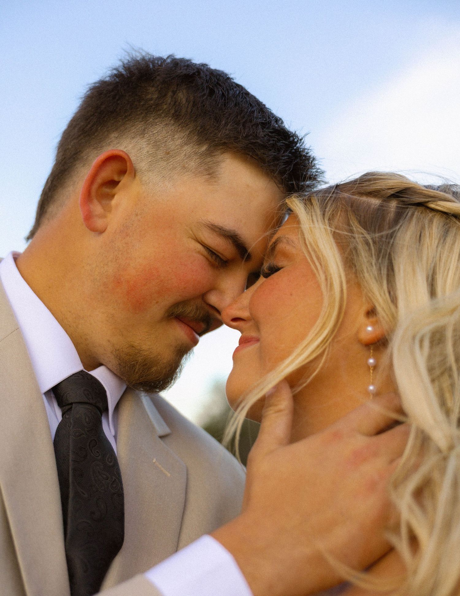 A couple in wedding attire share an intimate moment, touching foreheads and almost kissing outdoors.