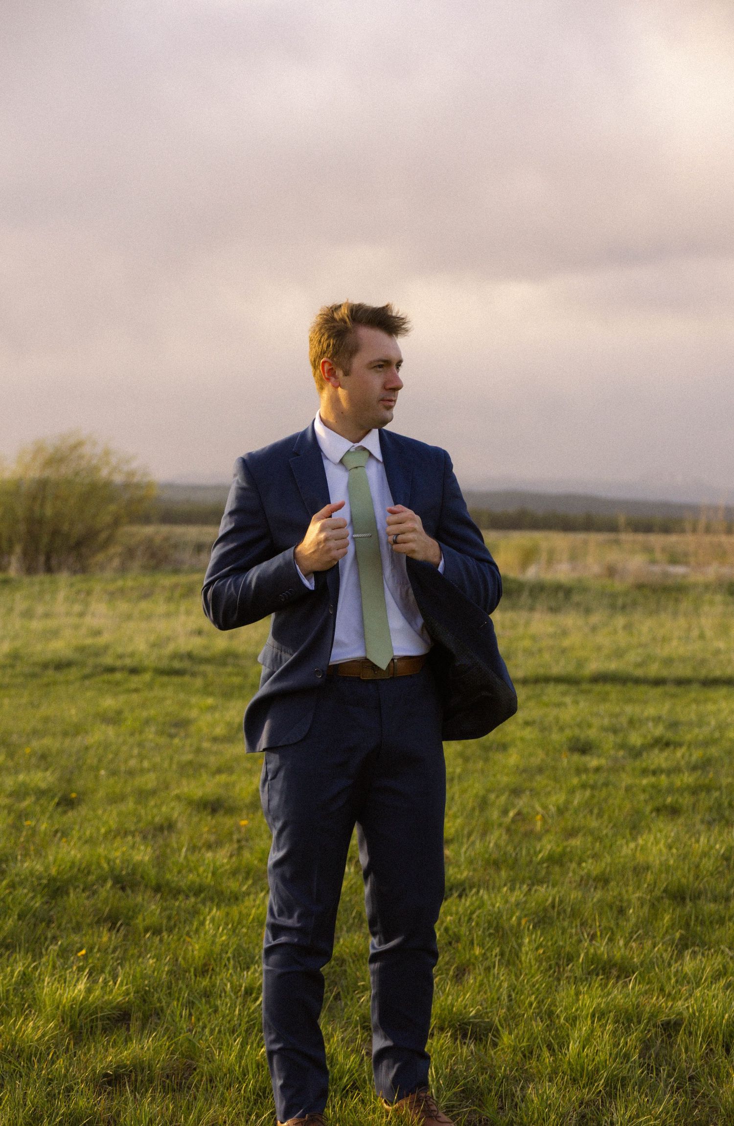 A young man in a navy blue suit and green tie stands confidently in a lush green field under a cloudy sky.