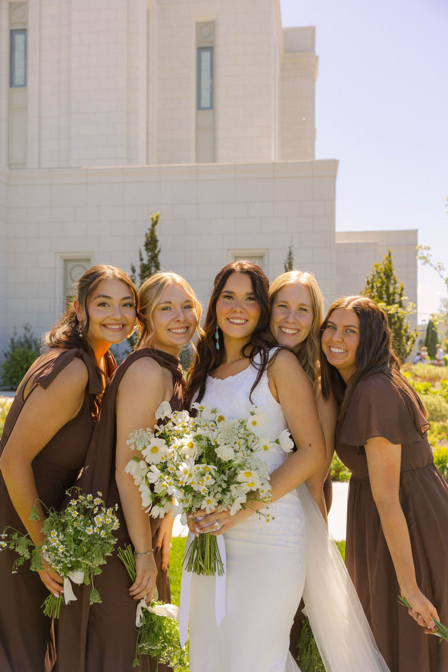 Bride in white gown with four bridesmaids in brown dresses holding white floral bouquets outside a temple.