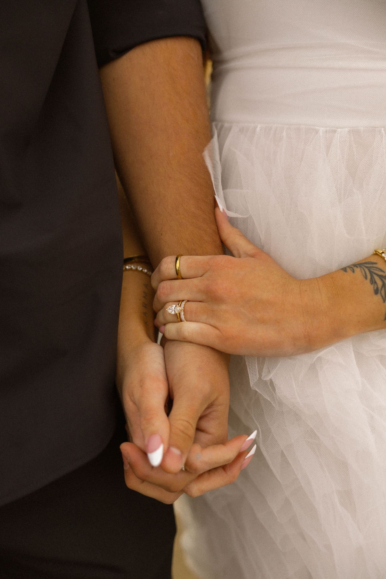 Close-up of bride and groom holding hands, showing wedding rings, bracelet, and tattoo, with white bridal gown.