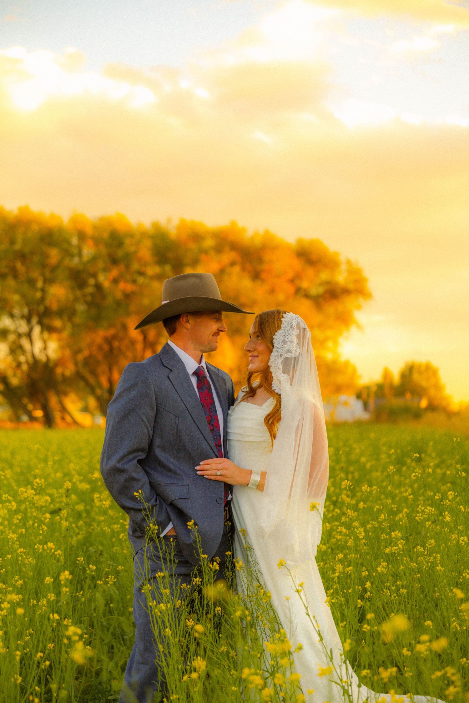 Bride and groom embrace in golden sunset field of yellow wildflowers, groom wearing cowboy hat and blue suit.
