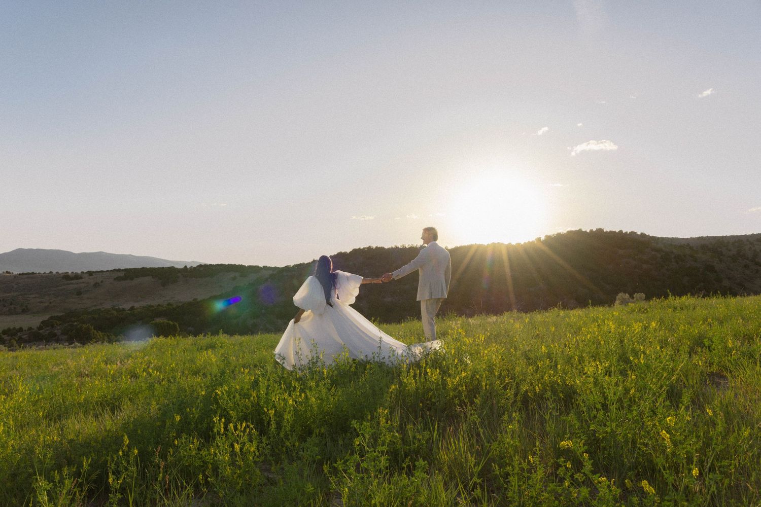 Bride and groom holding hands in a lush green meadow at golden sunset with rolling hills in the background.