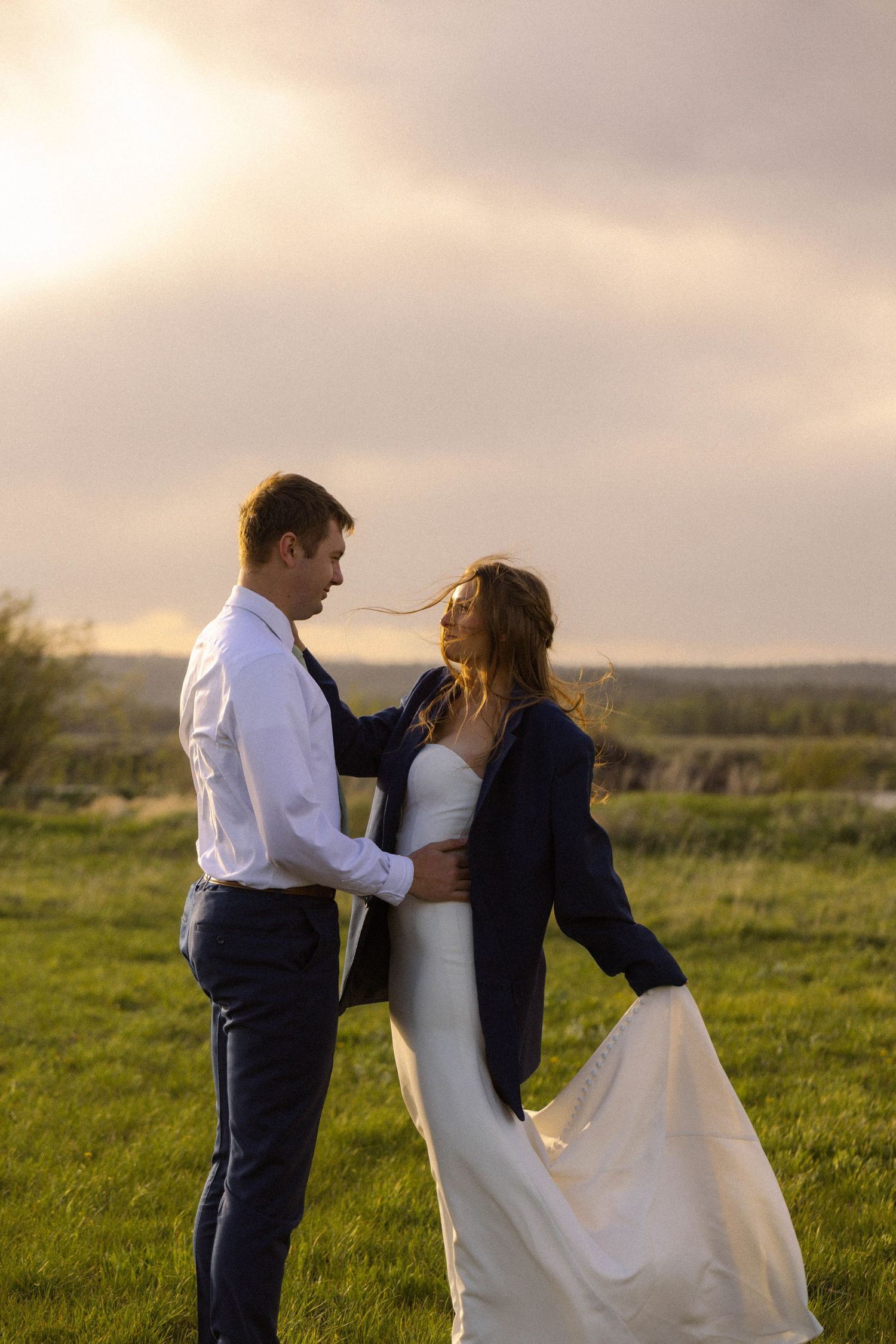 Bride and groom share a romantic sunset moment in a green field, she wearing his navy jacket over her wedding dress.