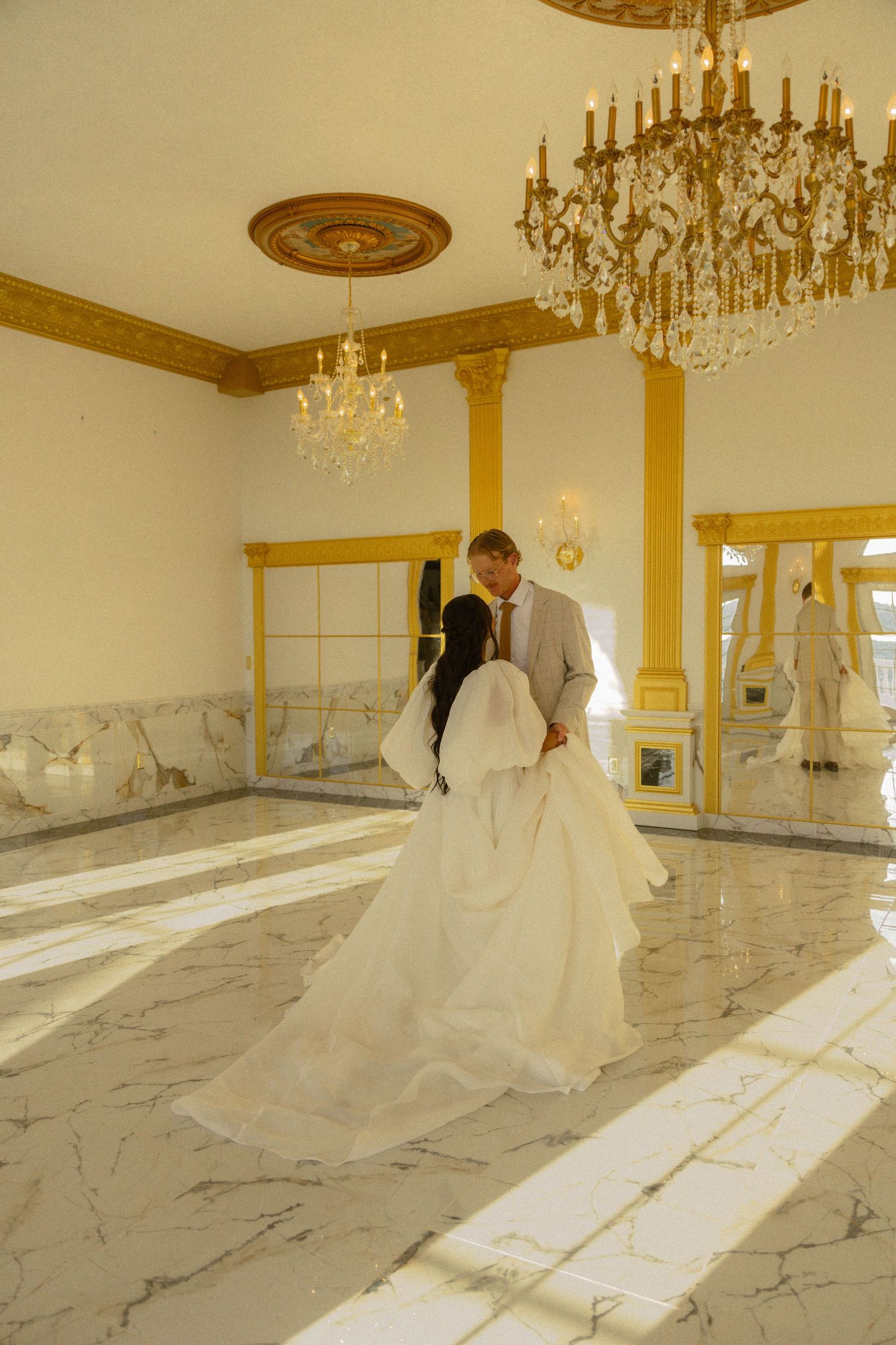 Bride and groom share first dance in elegant ballroom with marble floors and gold chandeliers.