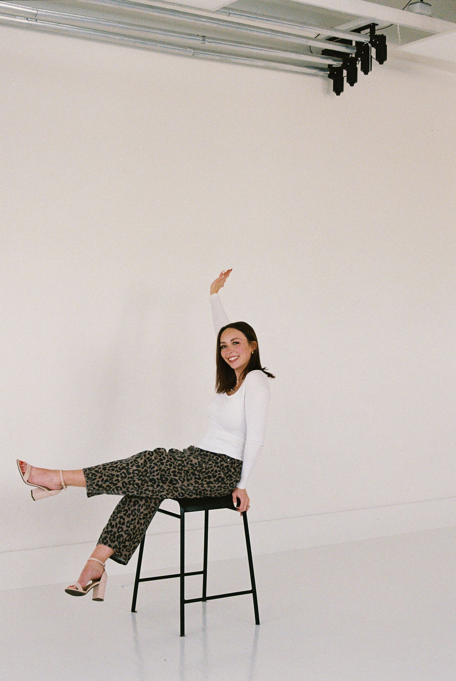 Woman in white top and leopard print pants sits on stool, raising one arm and smiling in bright studio.
