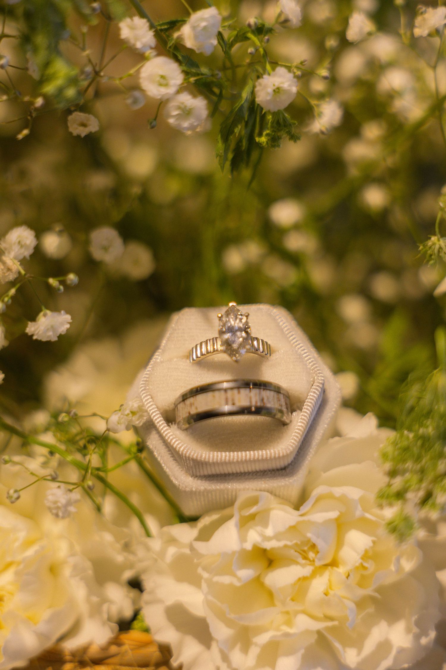 Two silver wedding rings in a hexagonal velvet box surrounded by white baby's breath and cream roses.