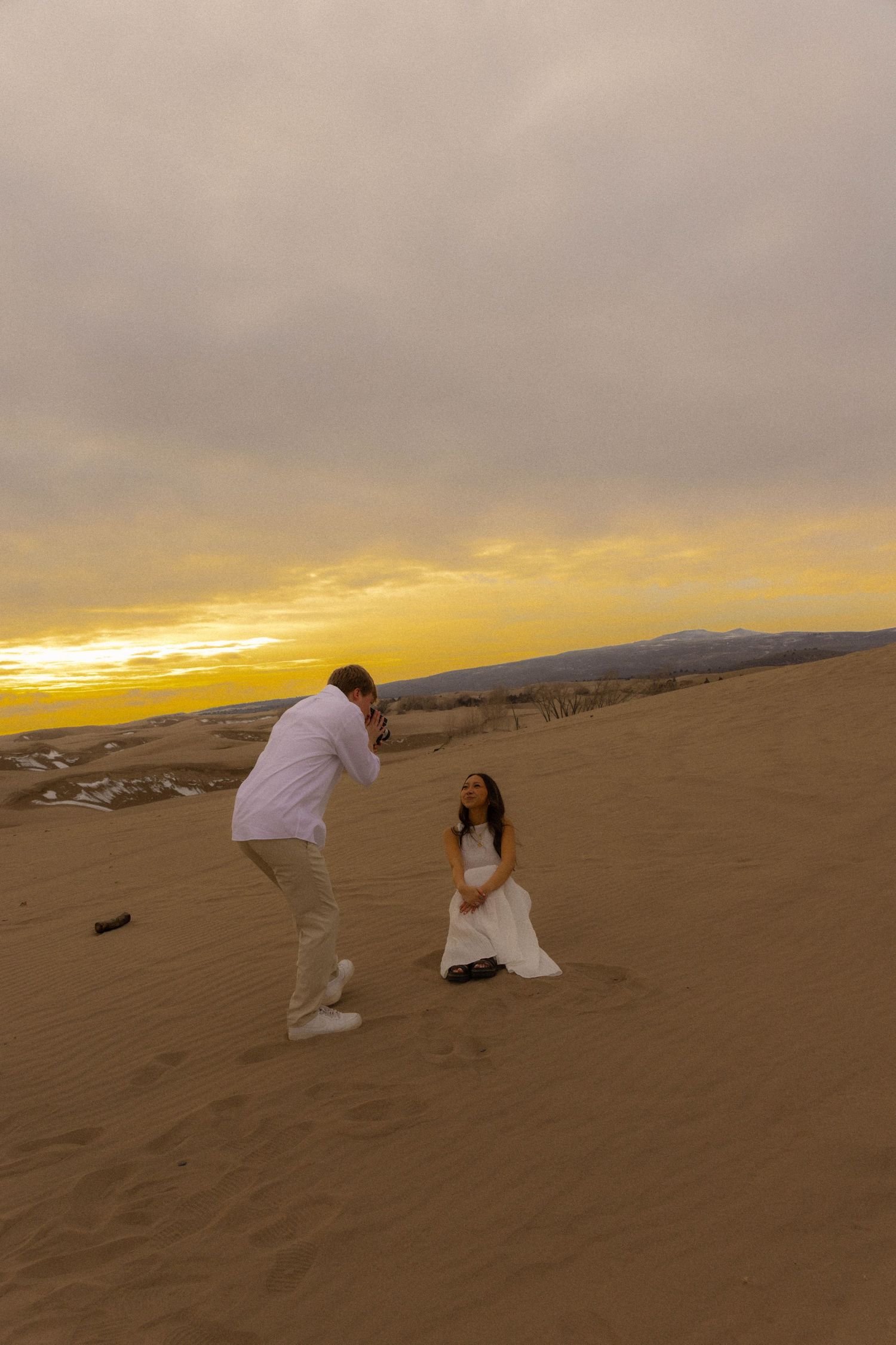 Man photographs woman in white dress on sand dunes at golden sunset.