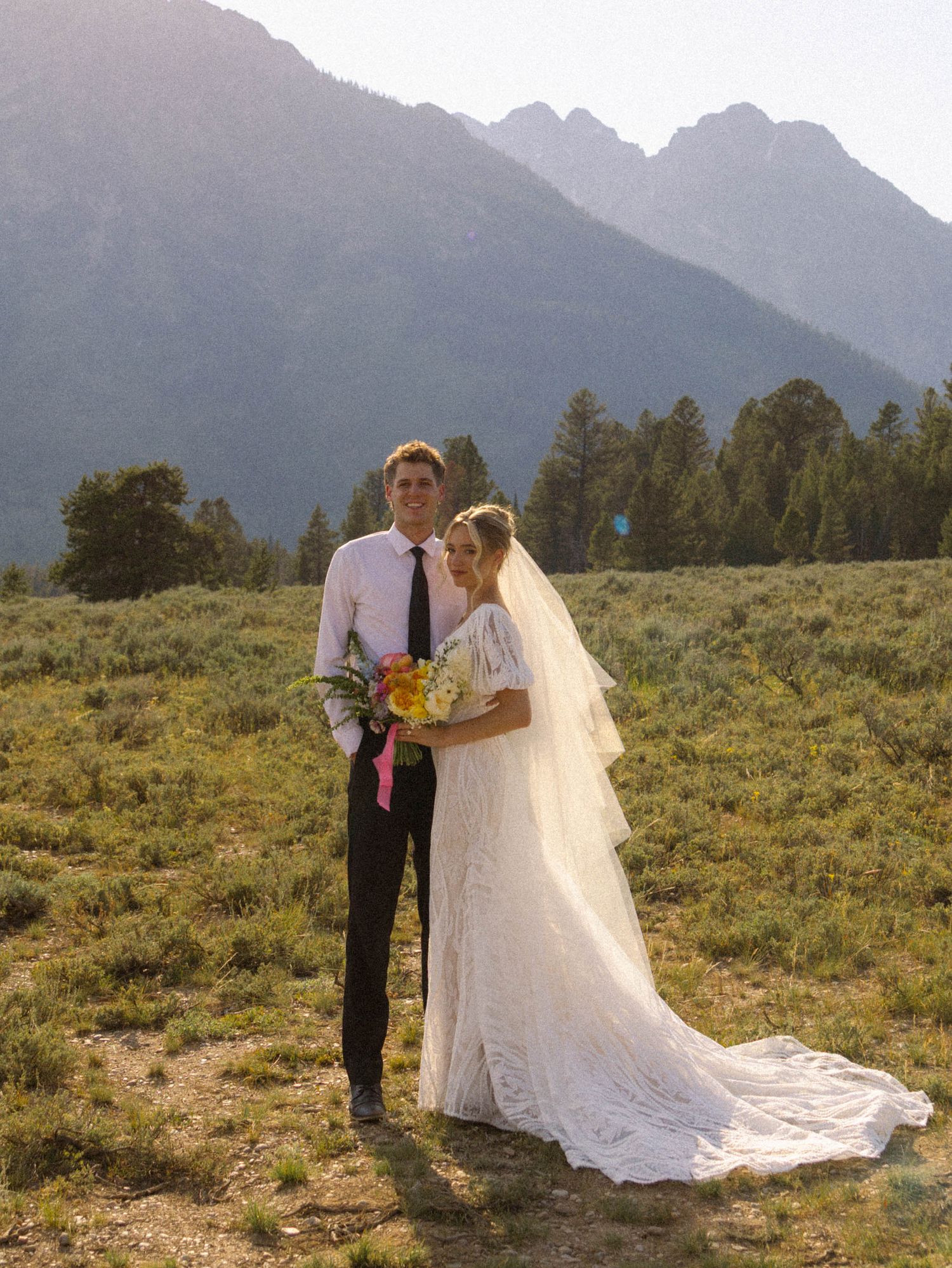 Bride and groom posing in mountain meadow, bride in lace gown with veil, groom in black suit with tie.