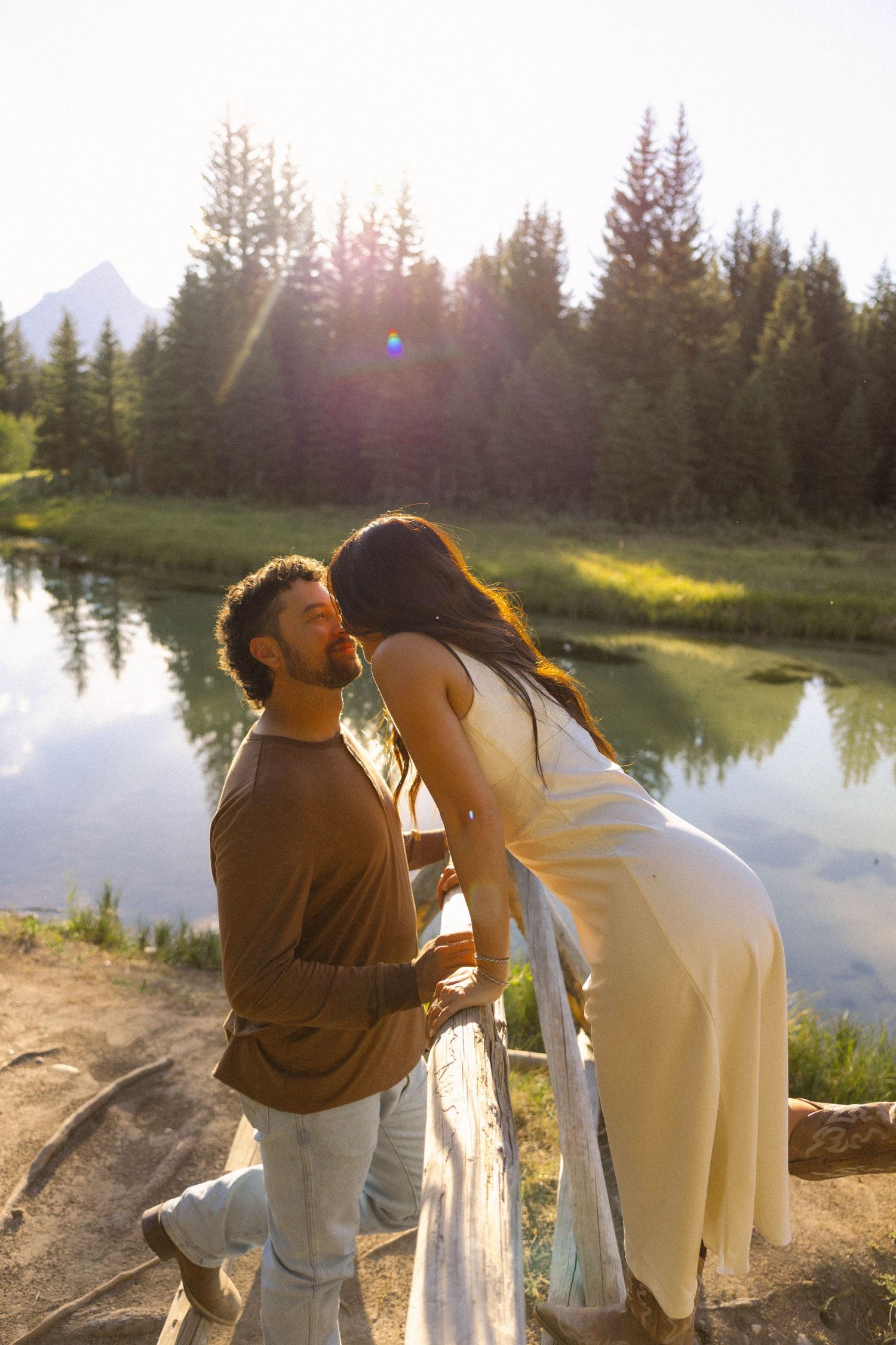Couple sharing a romantic moment by a serene mountain lake surrounded by pine trees in golden sunlight.