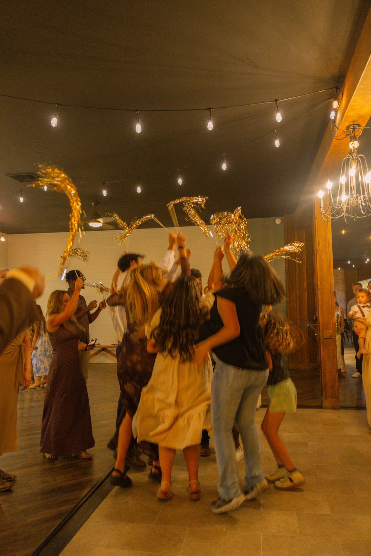 Group of women dancing and celebrating indoors under warm string lights at an evening party or wedding reception.