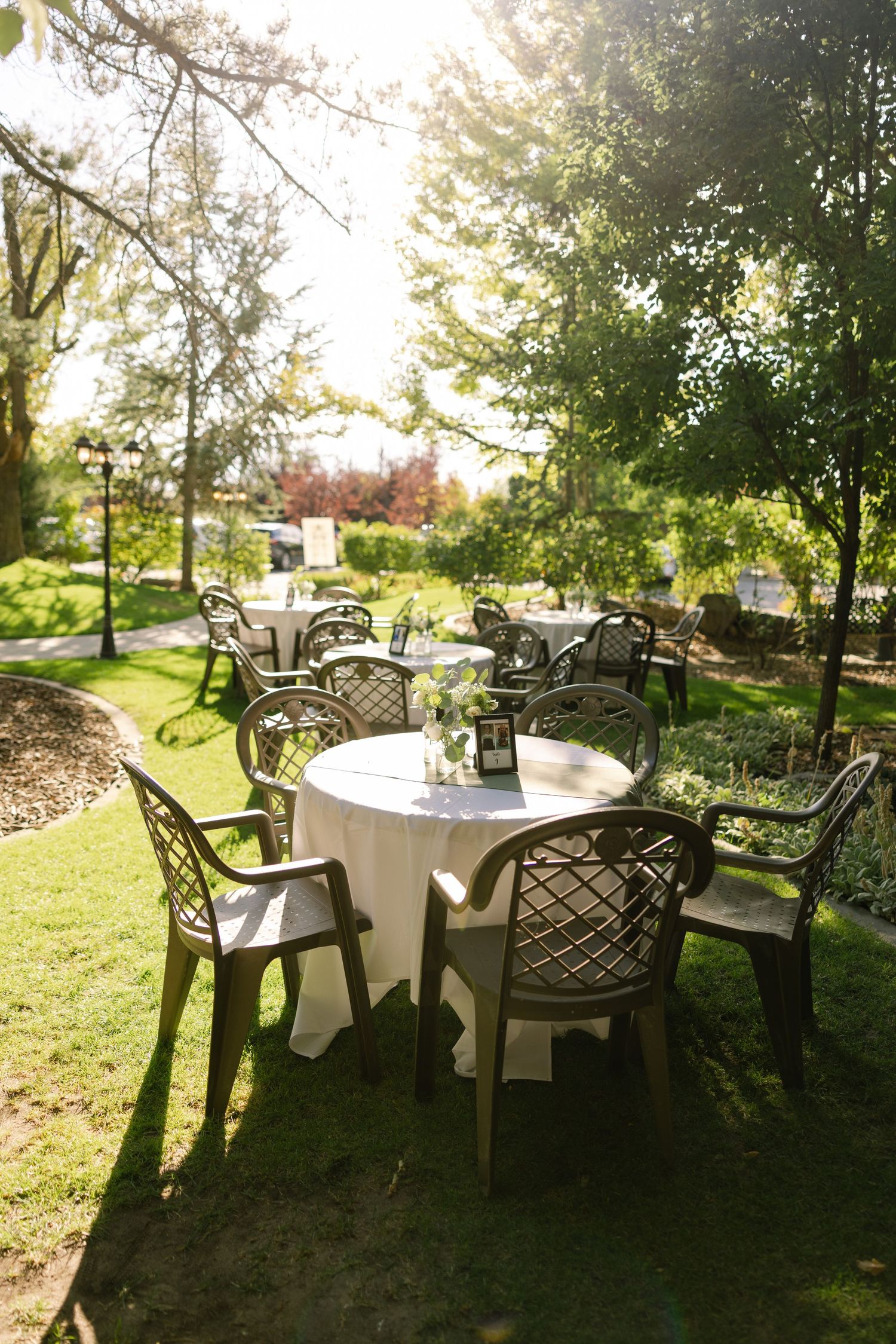 Outdoor garden dining setup with round tables, white tablecloths, and dark plastic chairs surrounded by lush green trees.