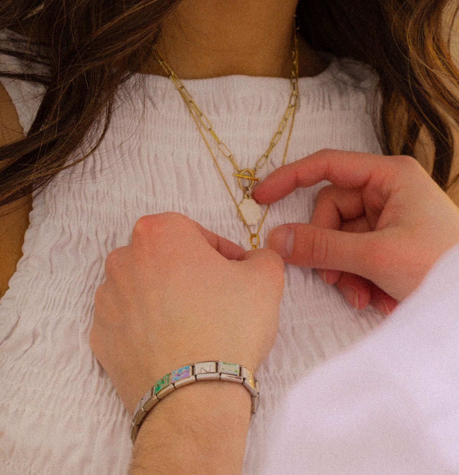 Woman wearing a gold chain necklace and silver bracelet with colorful stones, touching pendant on white top.